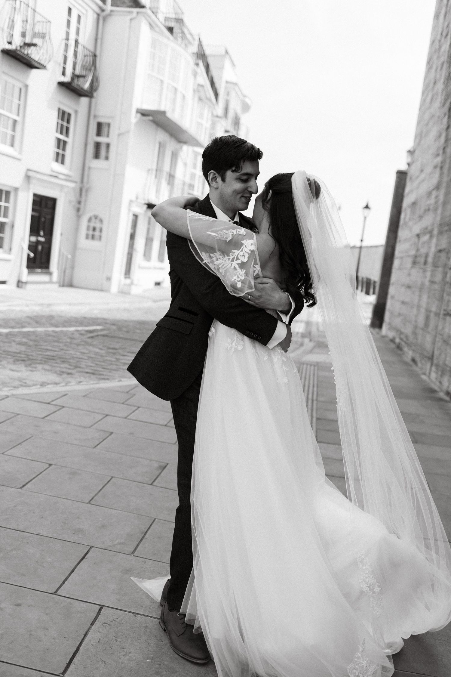 A black and white wedding photo of newlyweds embracing on a cobblestone street.