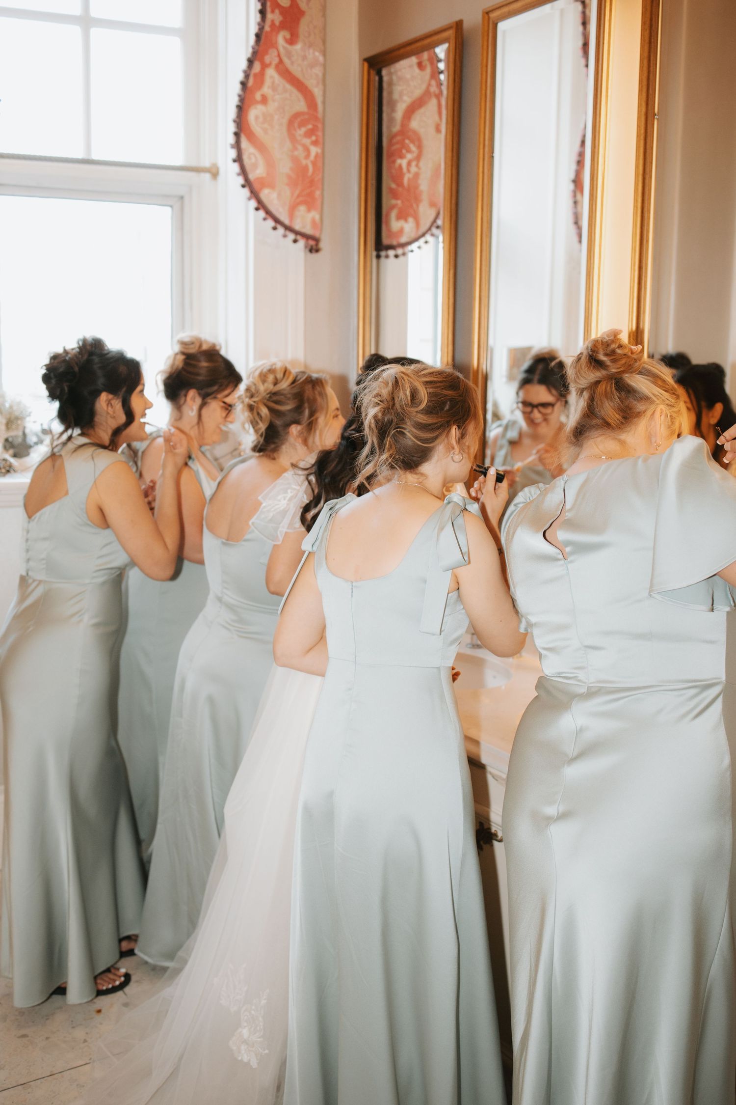 Bridesmaids in matching sage green dresses standing by a sunlit window.