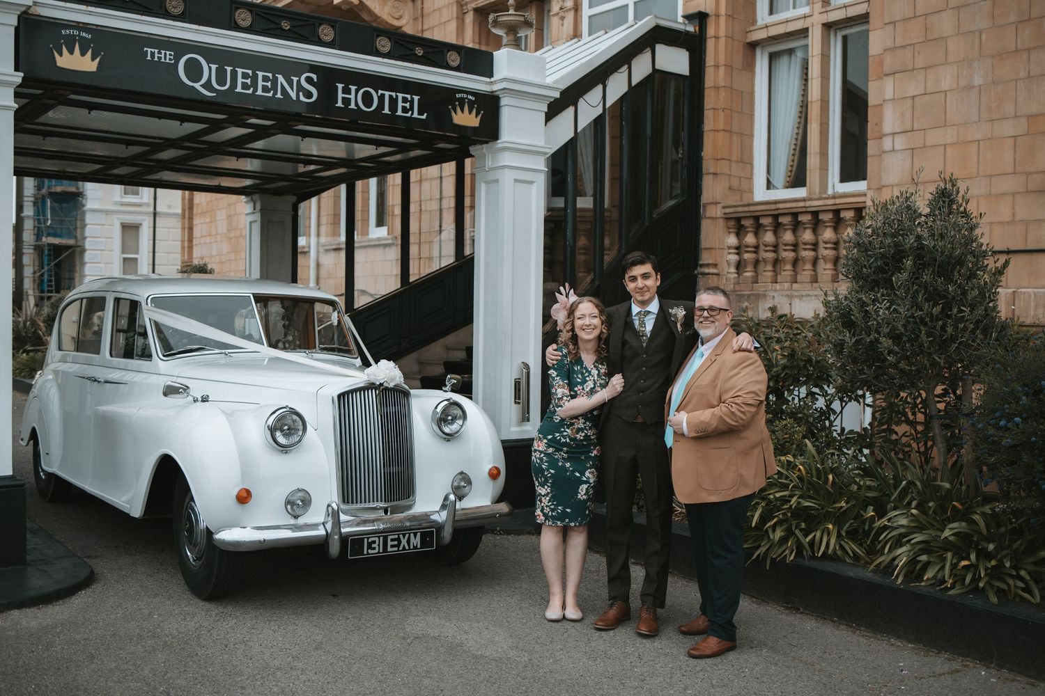 Exterior view of The Queens Hotel with classic architecture and vintage cars parked outside.