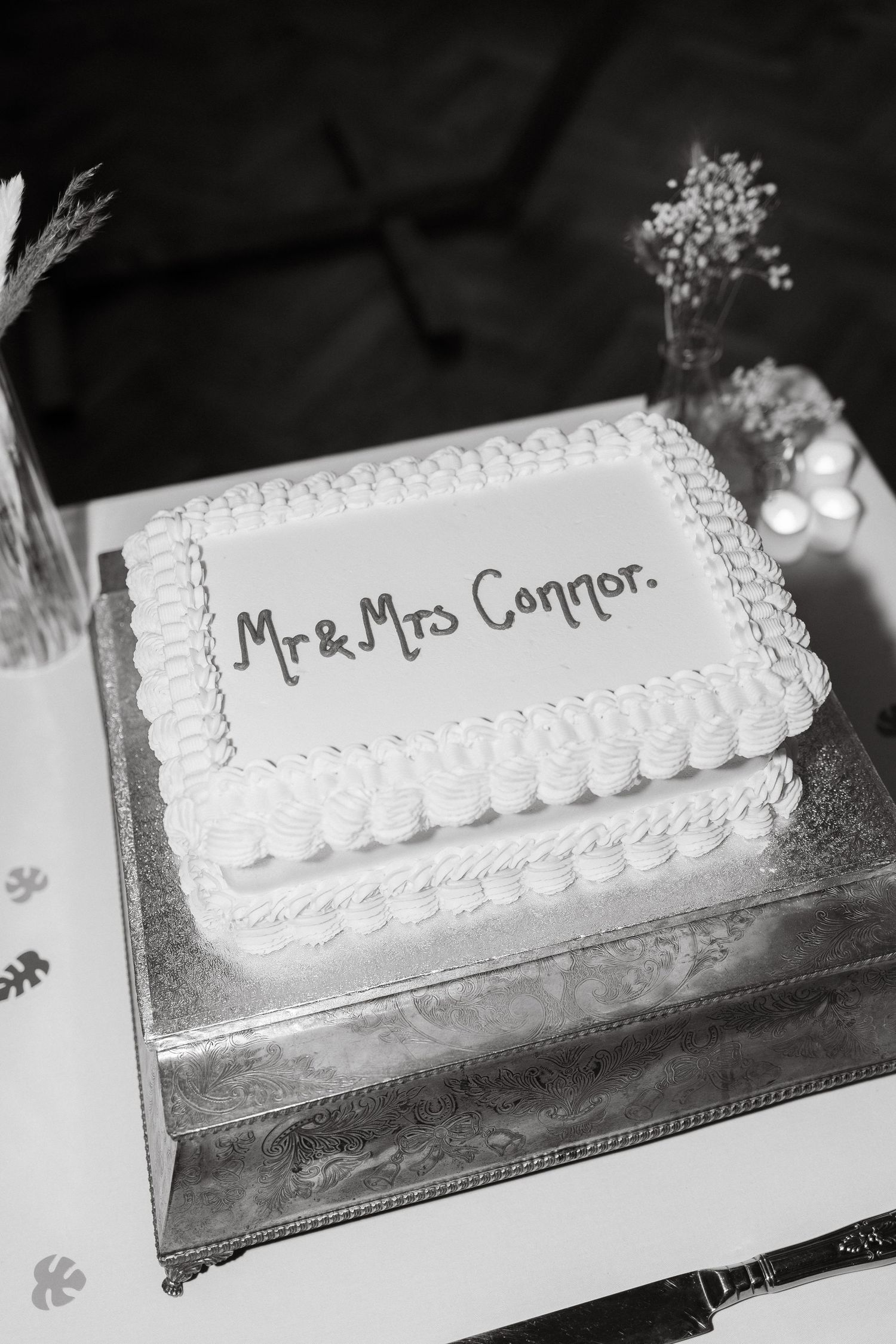 Black and white photo of a wedding cake with white frosting and decorative piping on a silver platter.