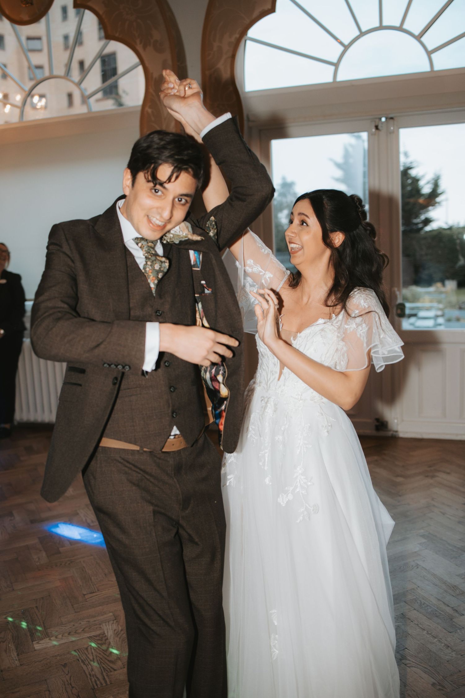 Couple dancing in a ballroom with arched windows and wooden floors during their first dance.