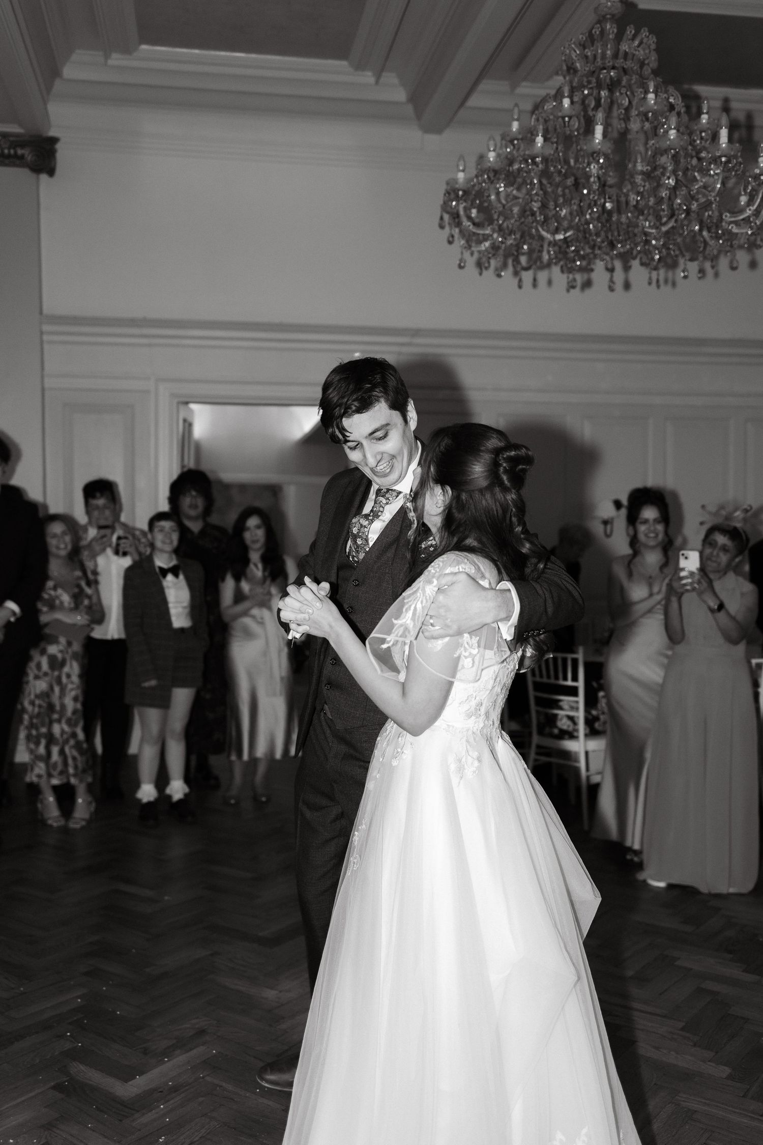 Couples dancing in an elegant ballroom with crystal chandeliers overhead.