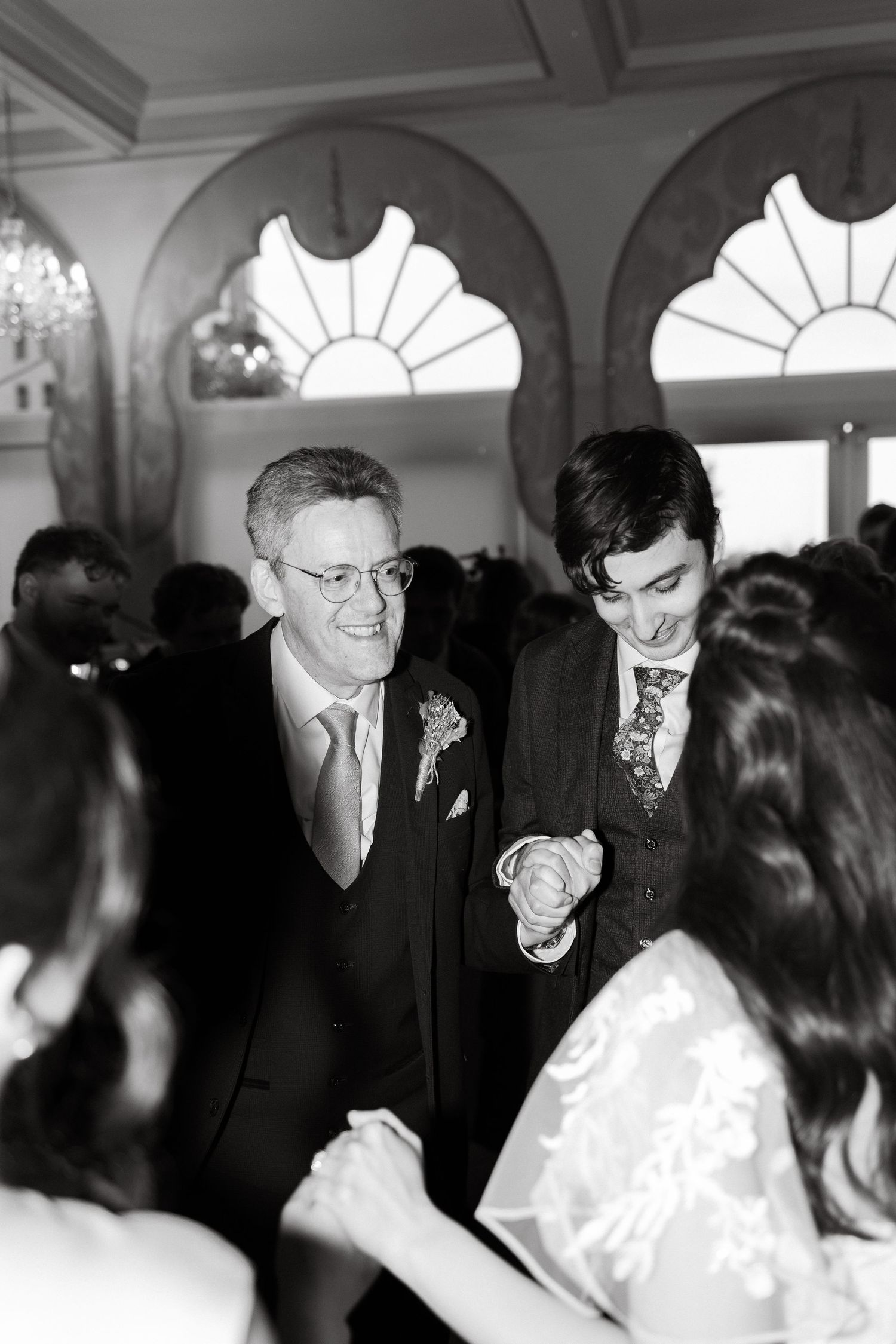 Wedding guests dancing and celebrating in an ornate room with arched windows.