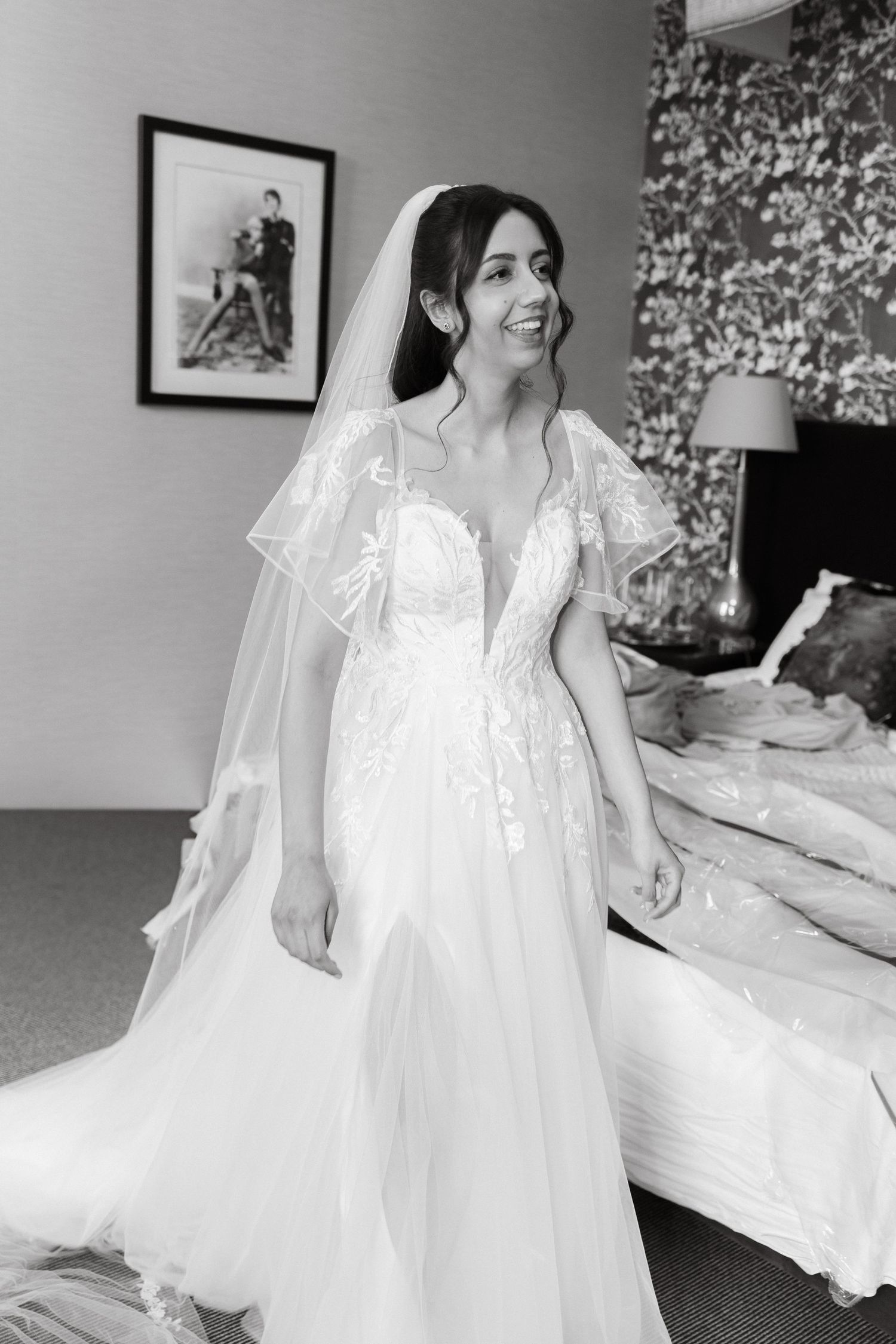 A bride in a delicate white lace gown with fluttering sleeves stands in an elegant bedroom setting.