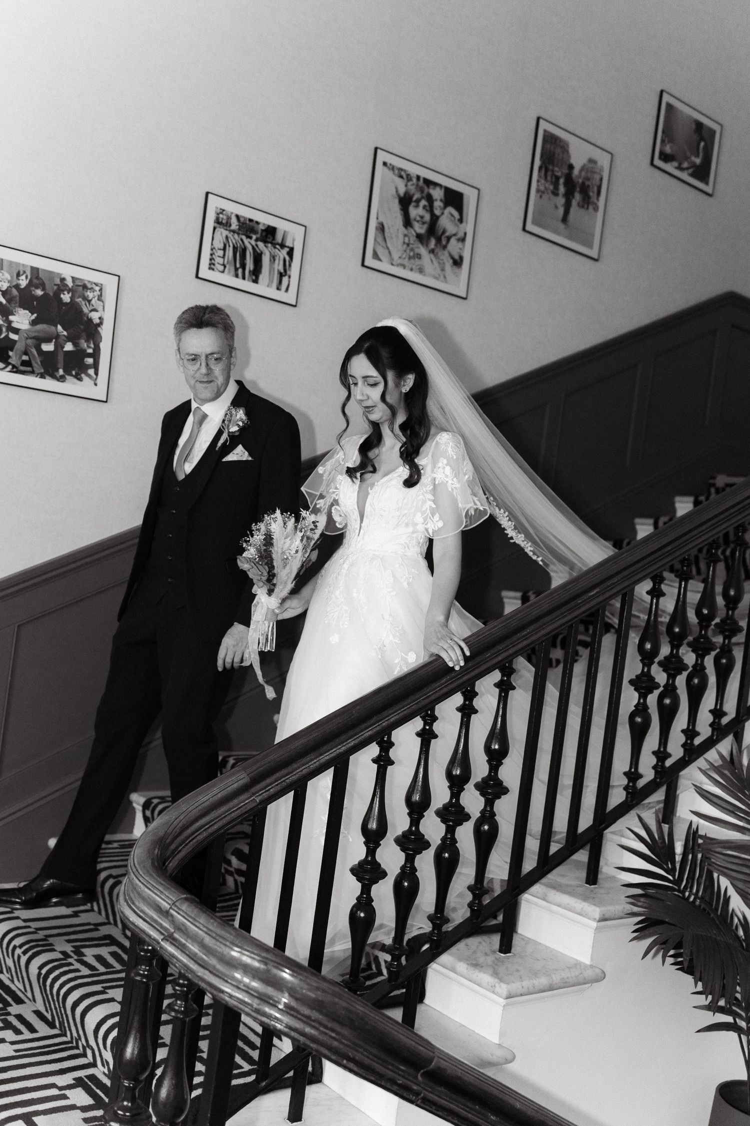 A wedding party descends a grand staircase with dark wooden banisters and framed photos on the wall.