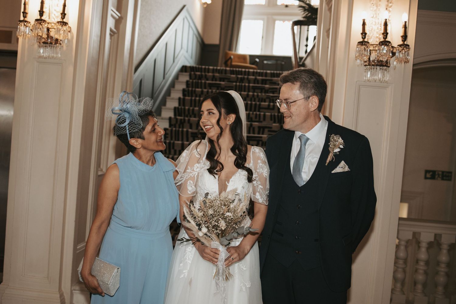 Wedding party members walk through an elegant white-walled hallway with chandeliers.