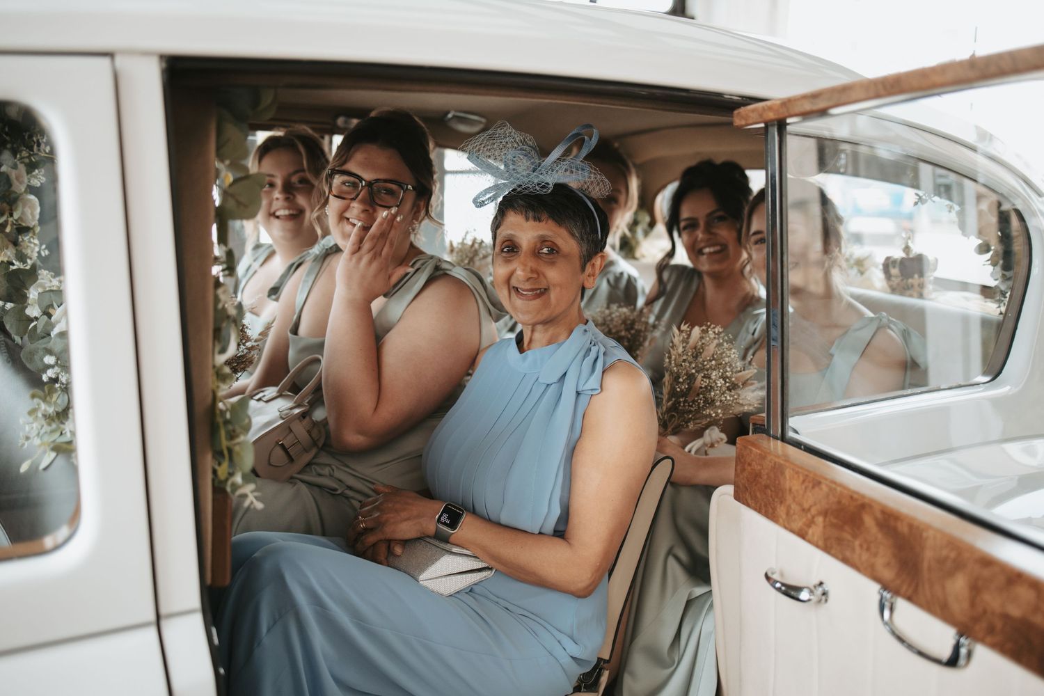 Wedding party members squeeze into a vintage white car with smiles and laughter.
