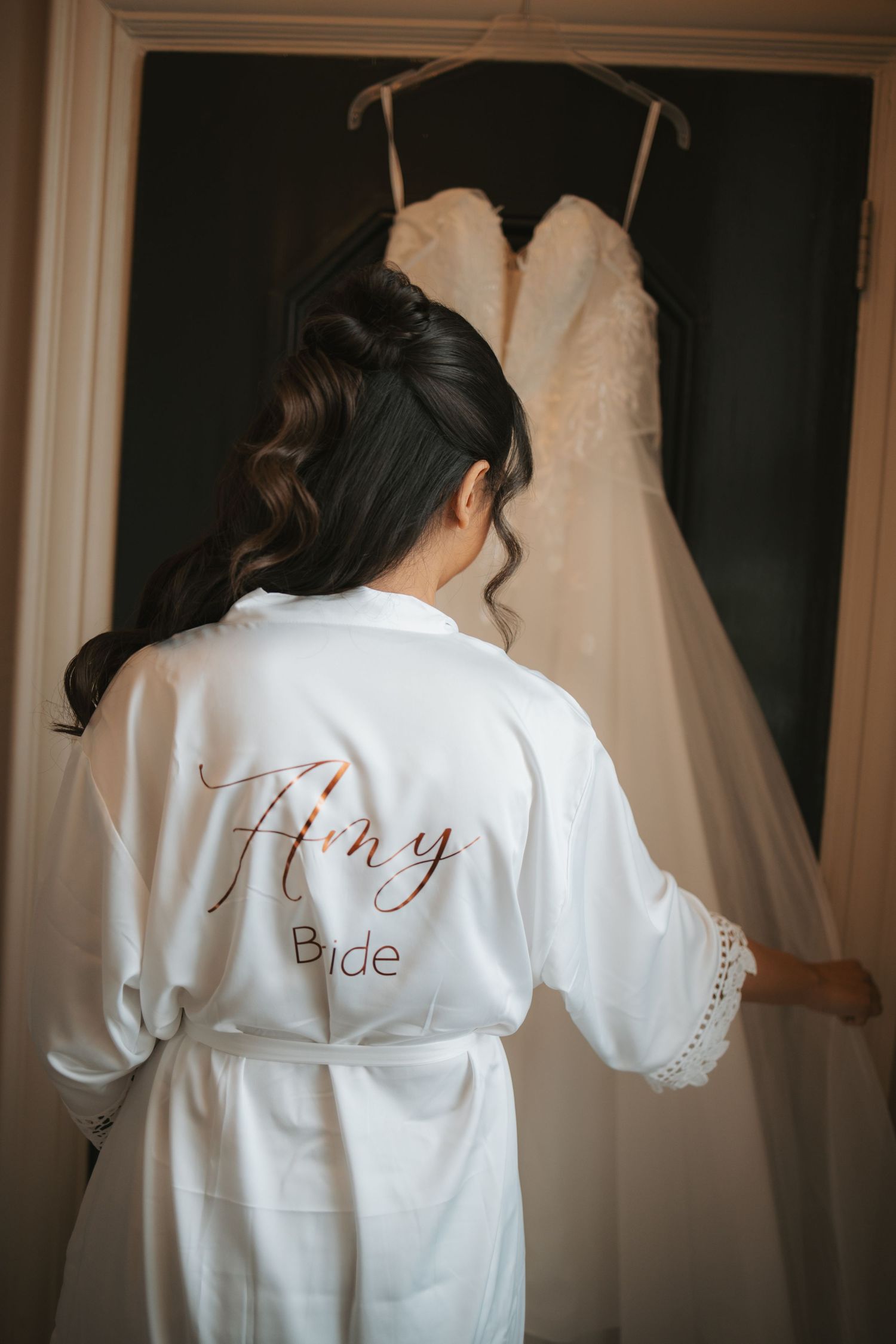 White bridal robe with gold lettering hanging beside wedding dress in preparation room.