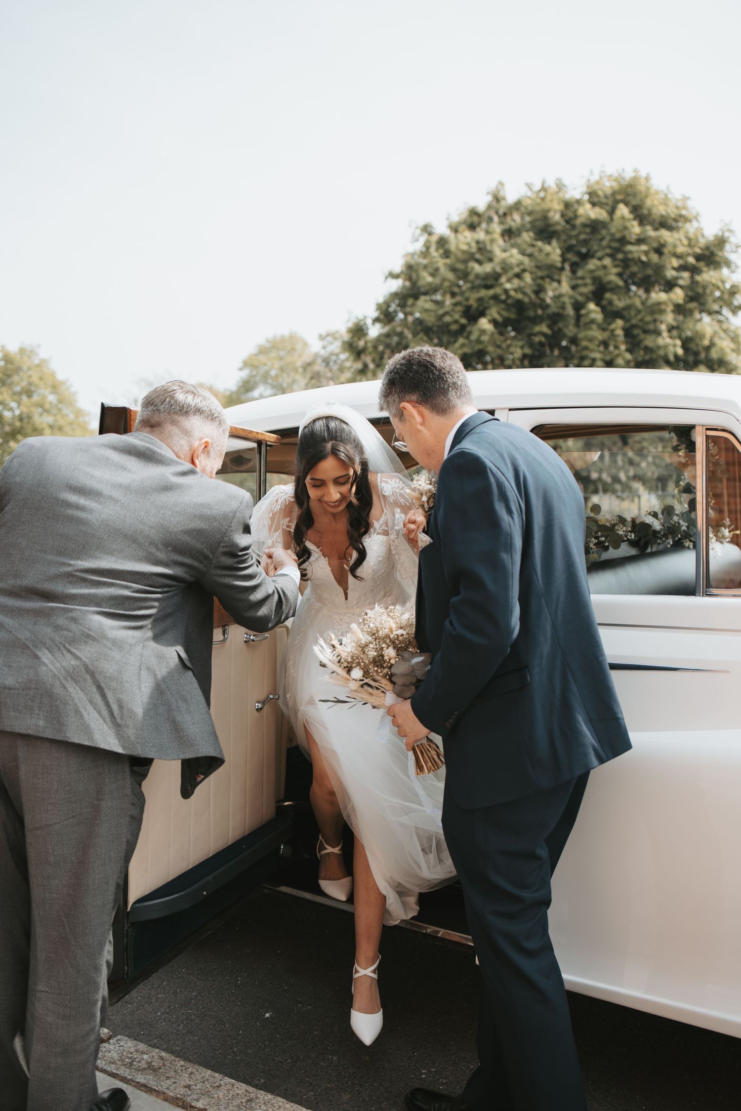 A classic white vintage car at a wedding as attendants help someone exit the vehicle.