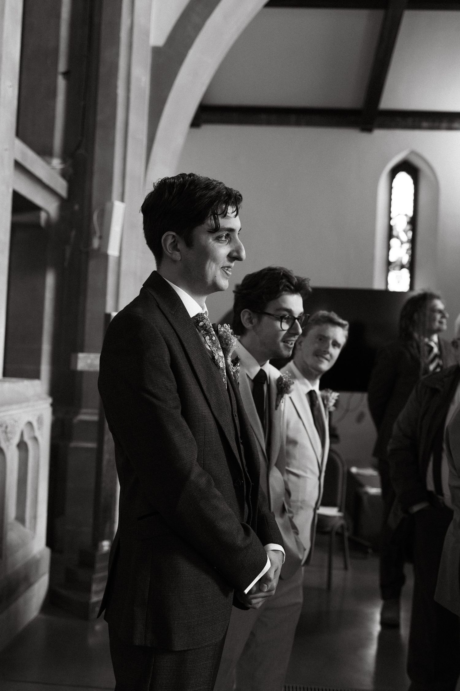 Wedding party members stand in a historic church with elegant arched windows and architectural details.