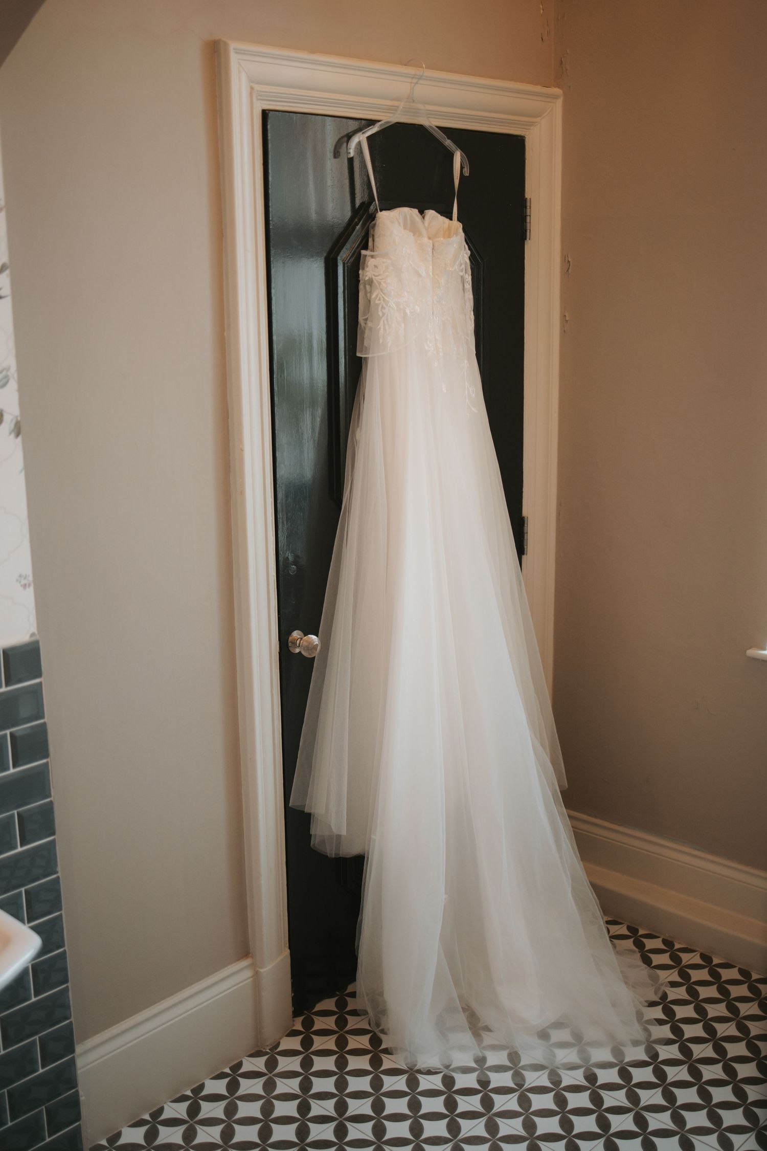 Elegant white wedding gown hanging in doorway against neutral wall and patterned floor.