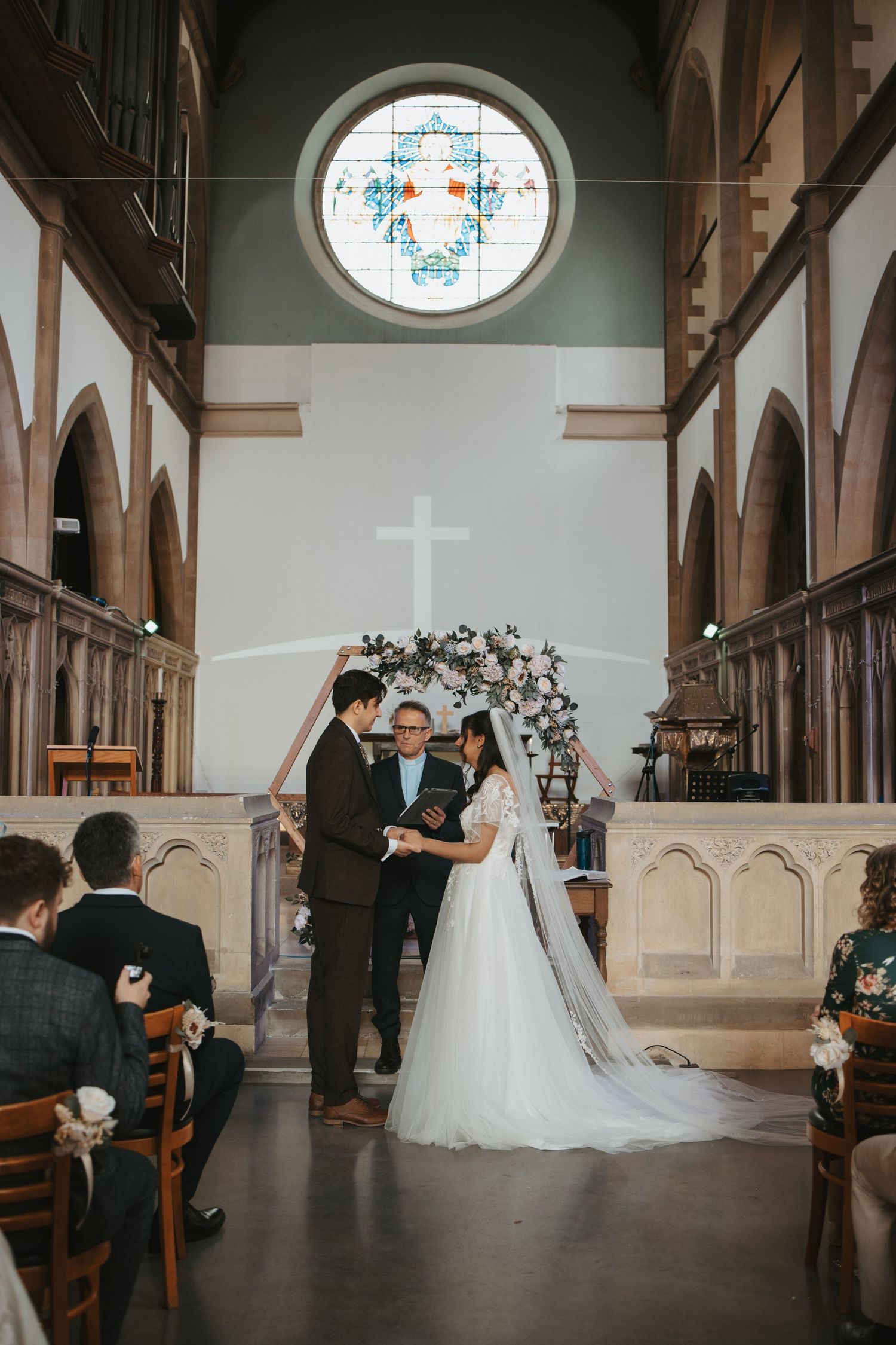 A wedding ceremony taking place under a stunning stained glass window and cross in a traditional church.
