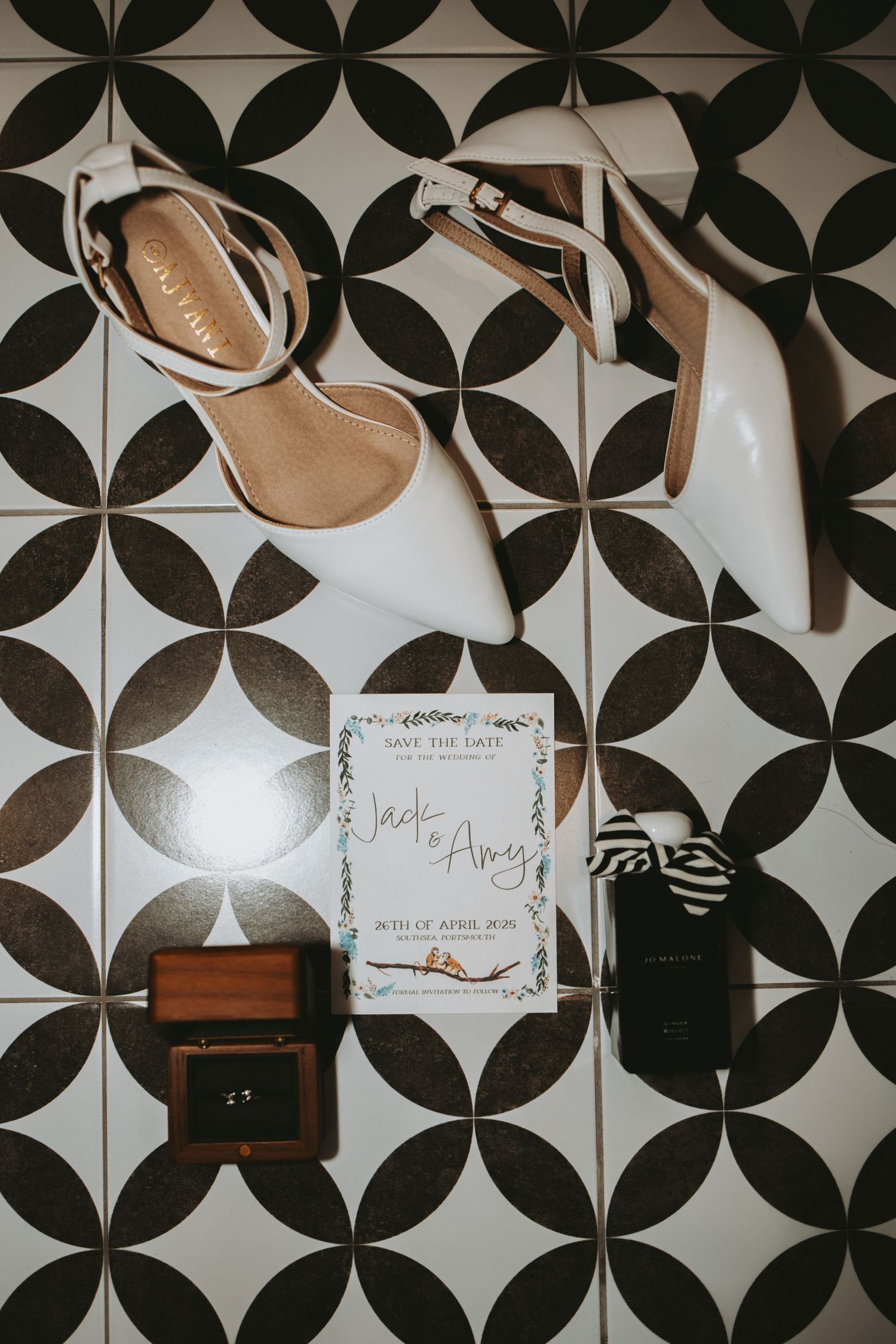 Wedding details flatlay with white shoes, invitation and ring box on geometric patterned tile.