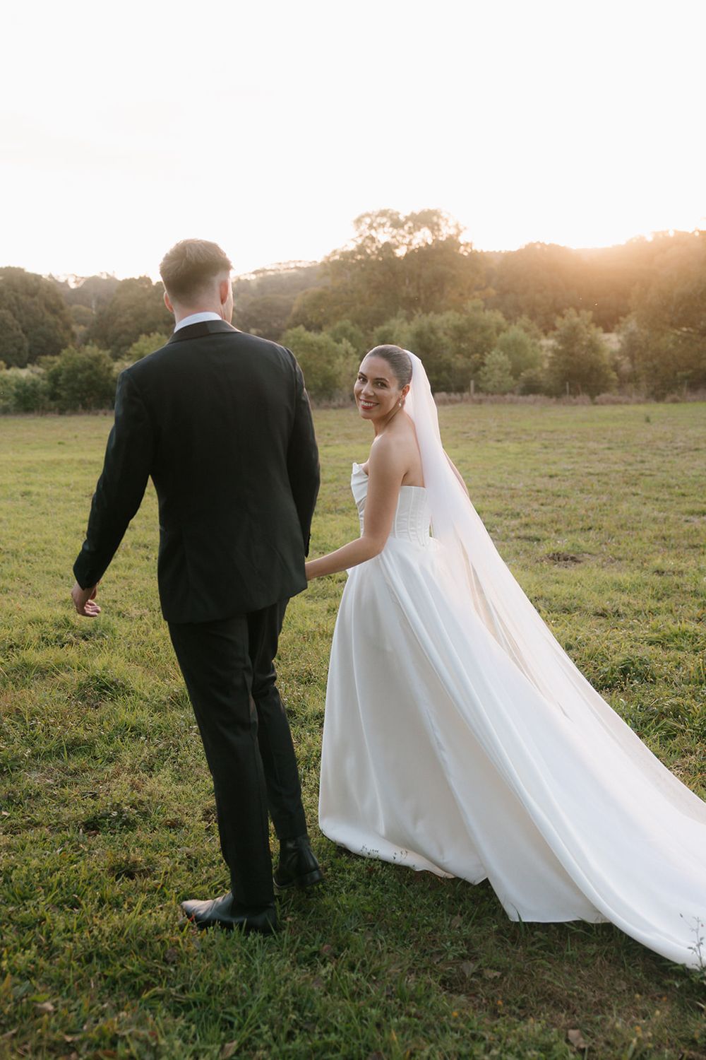 A couple in wedding attire walks through a sunlit field at golden hour as the bride's veil flows in the breeze.