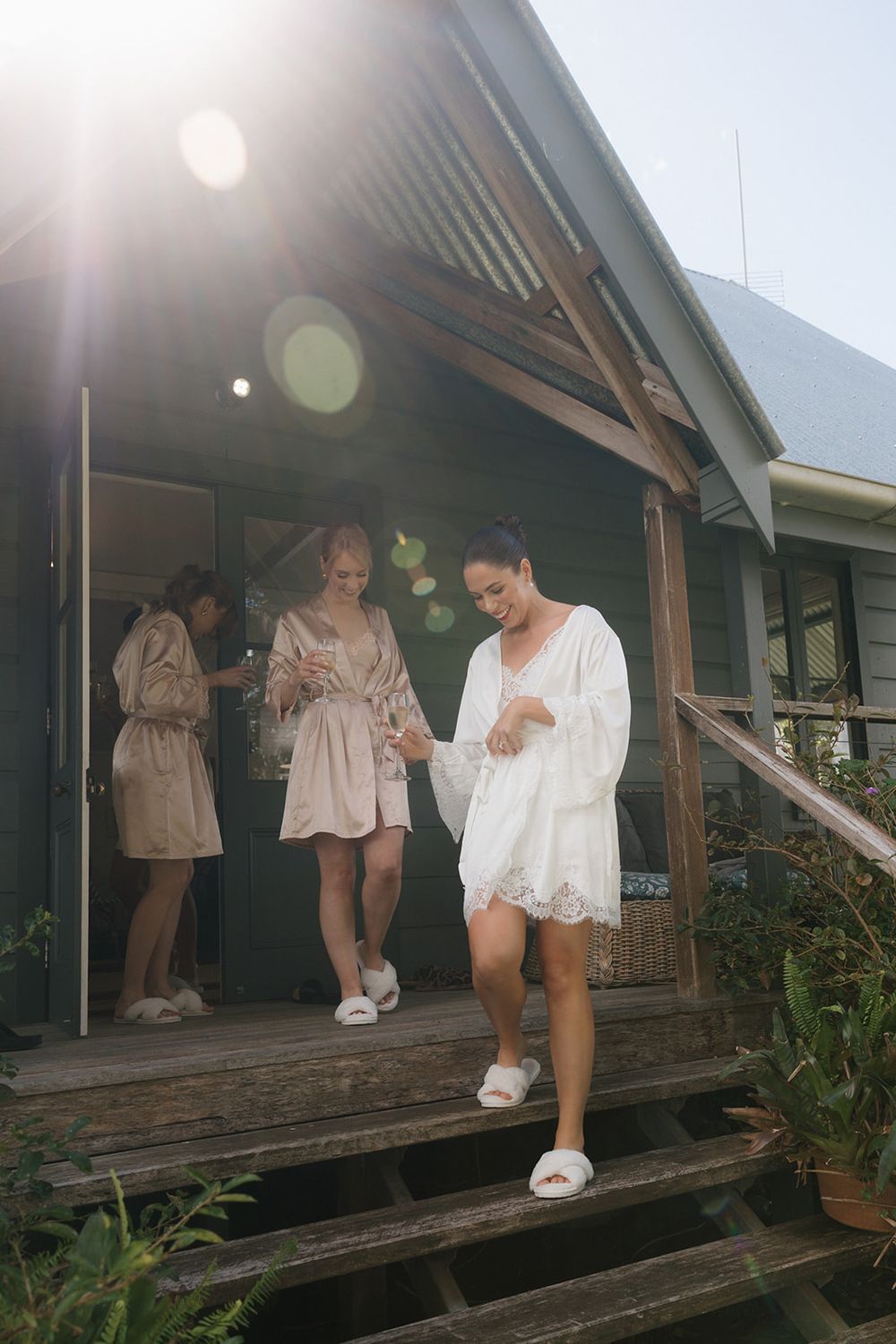 People in white outfits stand on a rustic wooden deck of a dark-colored cabin on a sunny day.