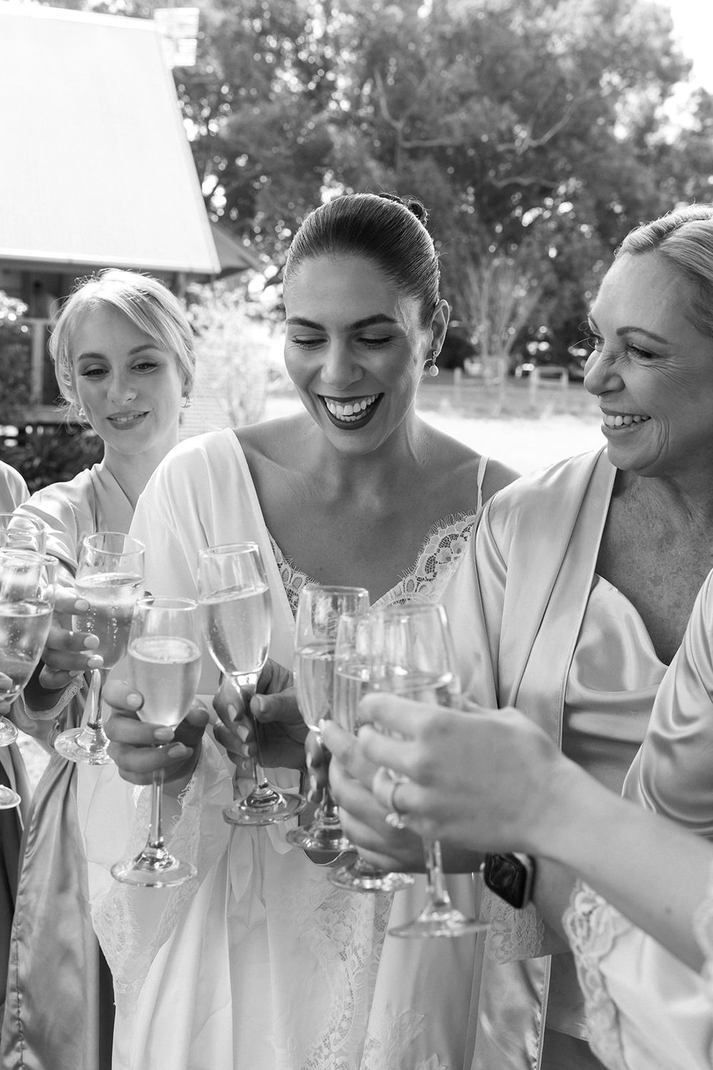 A group of bridesmaids in white dresses laugh and share drinks together during a wedding celebration.