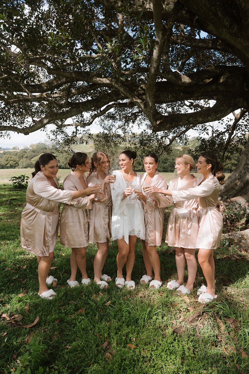 A group wearing matching neutral-toned outfits poses together under a large tree in a pastoral outdoor setting.