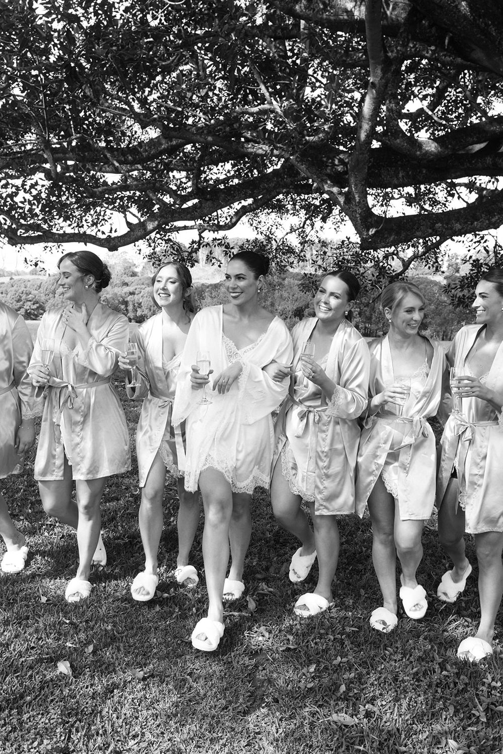 A group of people in white outfits sit in a row beneath a large tree branch in a black and white photograph.