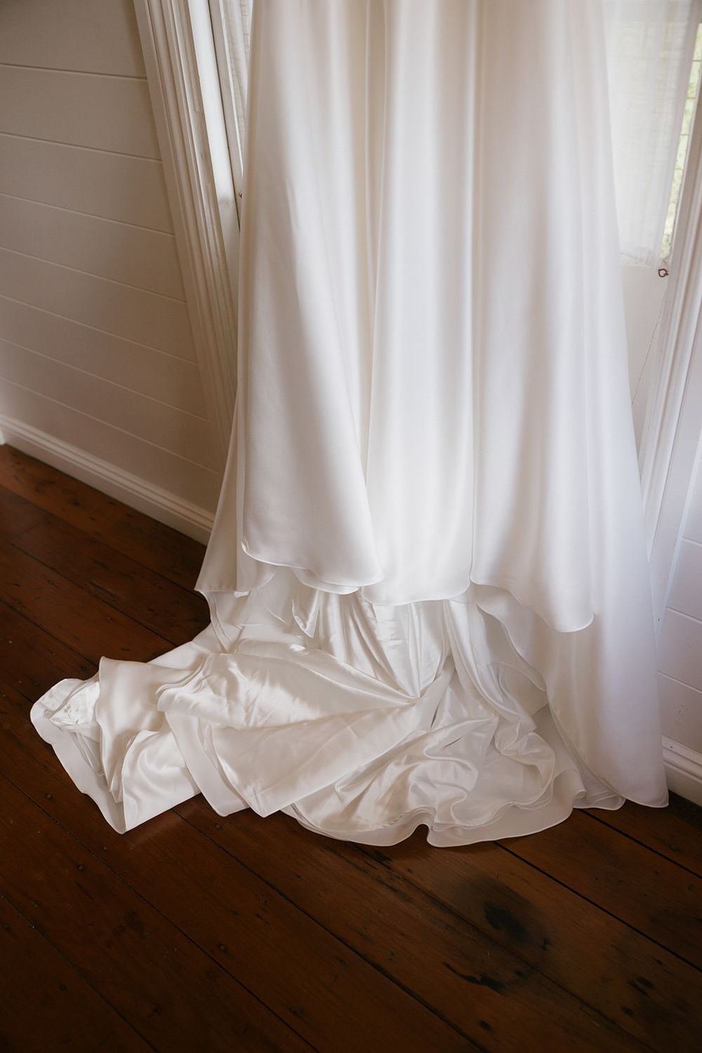 Close-up view of white wedding dress hem cascading across dark hardwood flooring in elegant folds.
