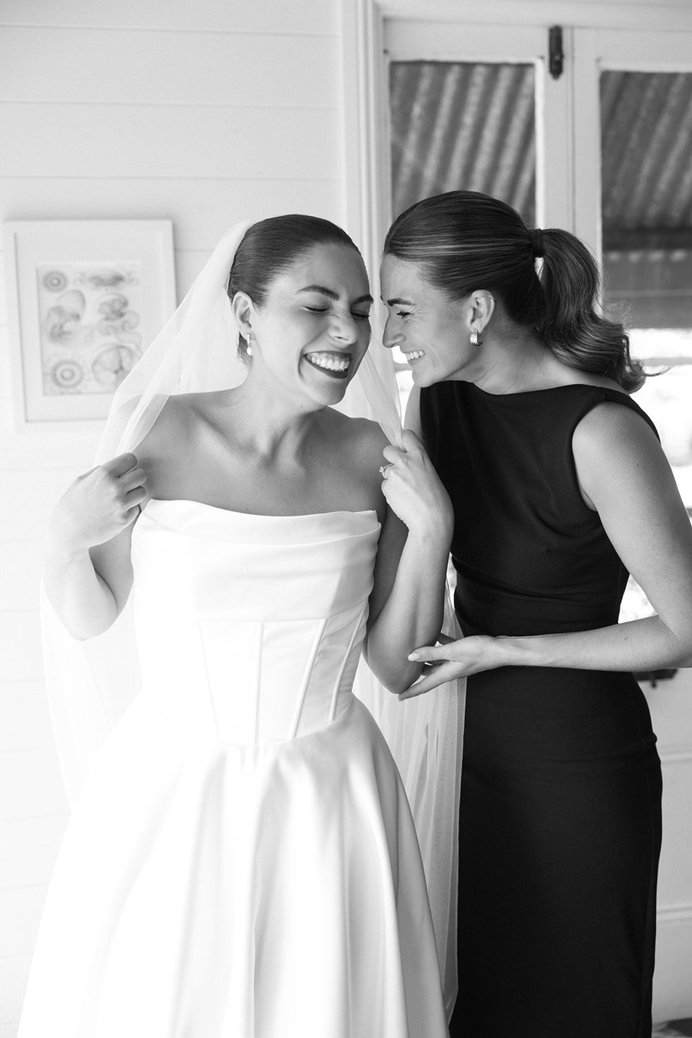 Black and white photo showing bridesmaids in dark dresses helping a bride get dressed for her wedding.