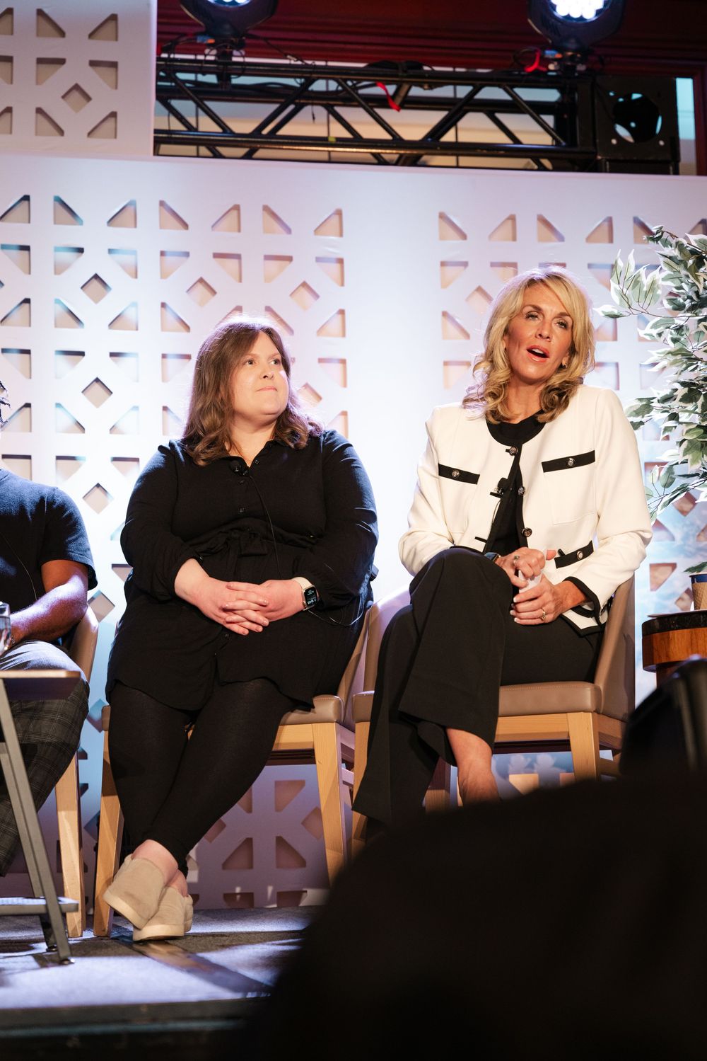 Two women sit on stage during a panel discussion against a white decorative backdrop with geometric patterns.