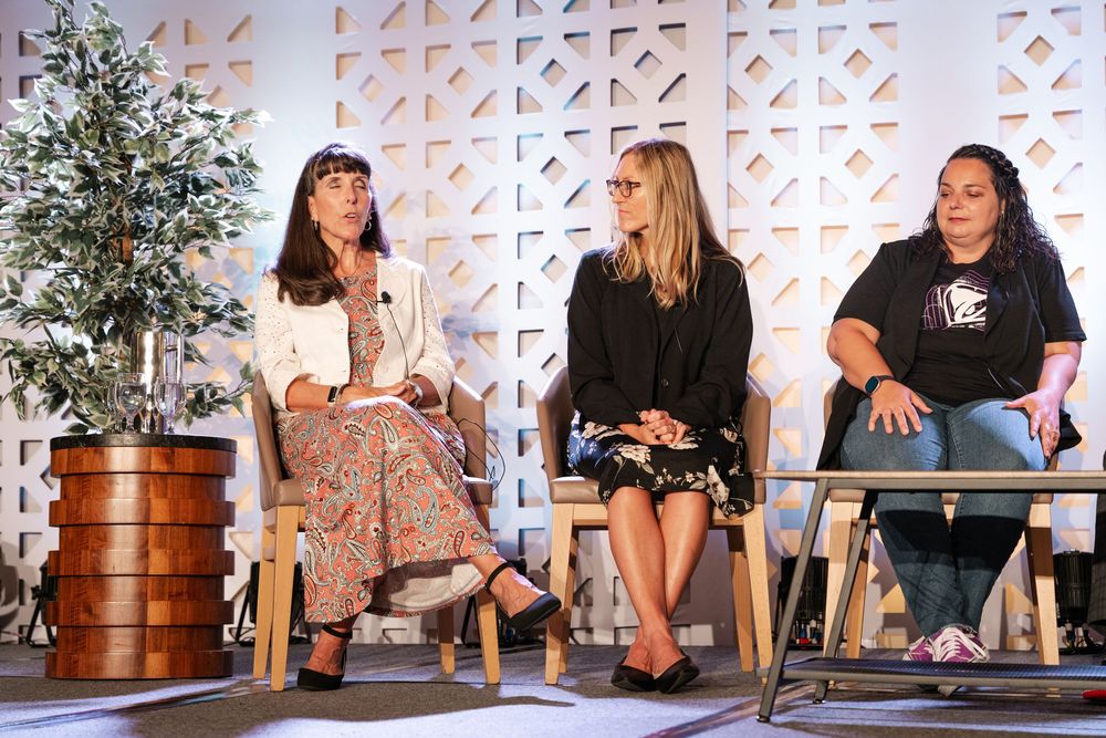 A group of speakers sit on chairs during a panel discussion with a decorative geometric wall pattern in the background.