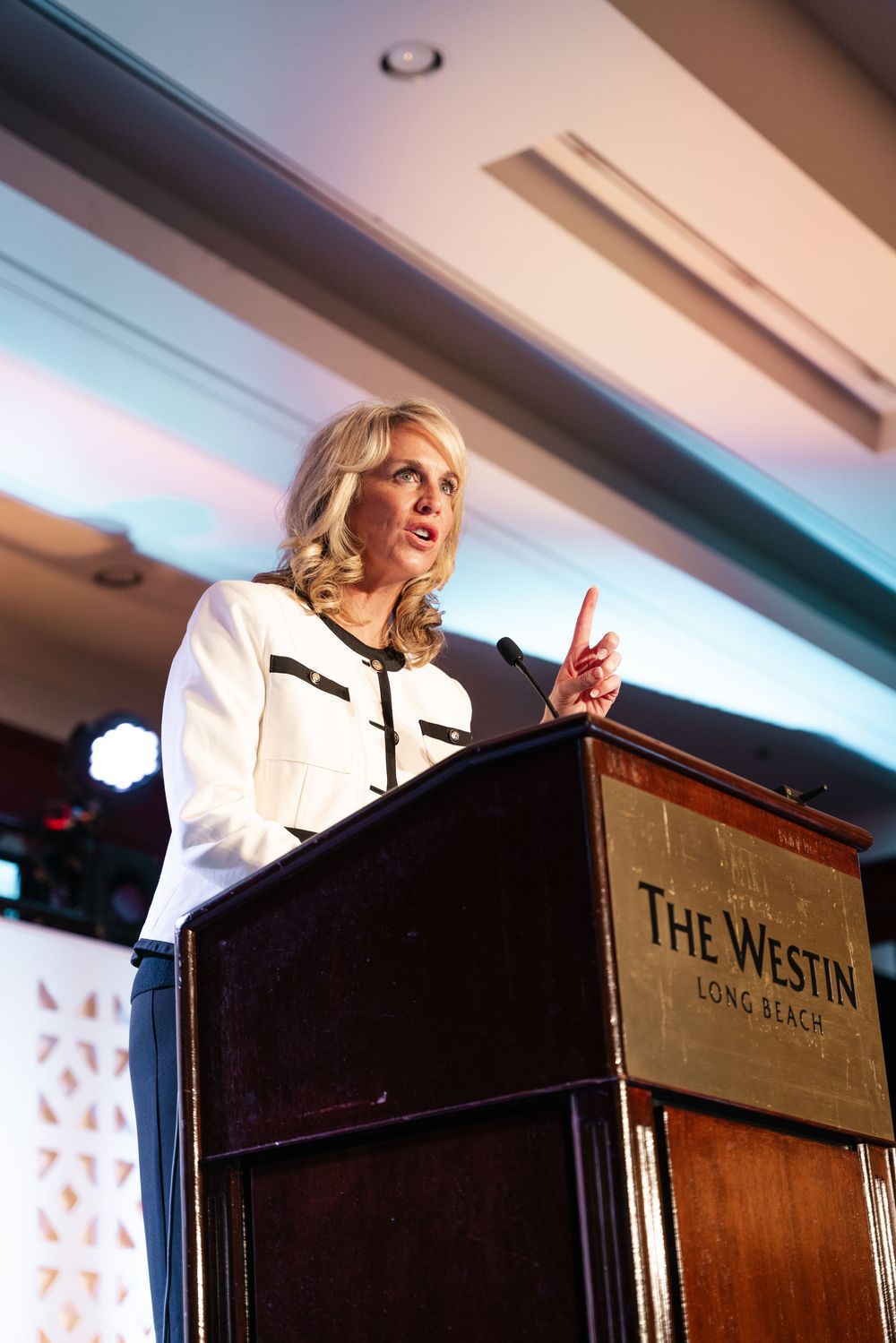 A speaker in a white blazer gestures animatedly while presenting at a wooden podium with The Westin branding.