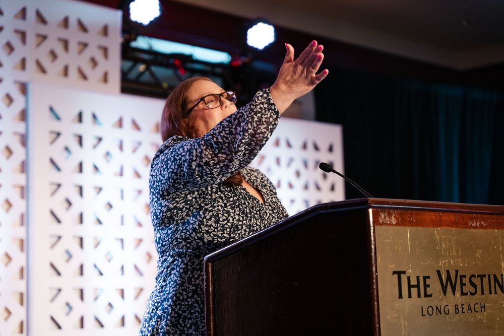 Speaker in a patterned dress gestures enthusiastically at podium during presentation at The Westin conference venue.