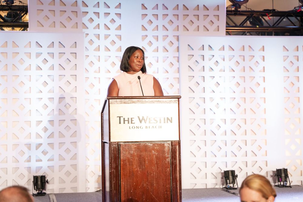 A speaker stands at a wooden podium on stage with a geometric patterned backdrop at a conference or event.
