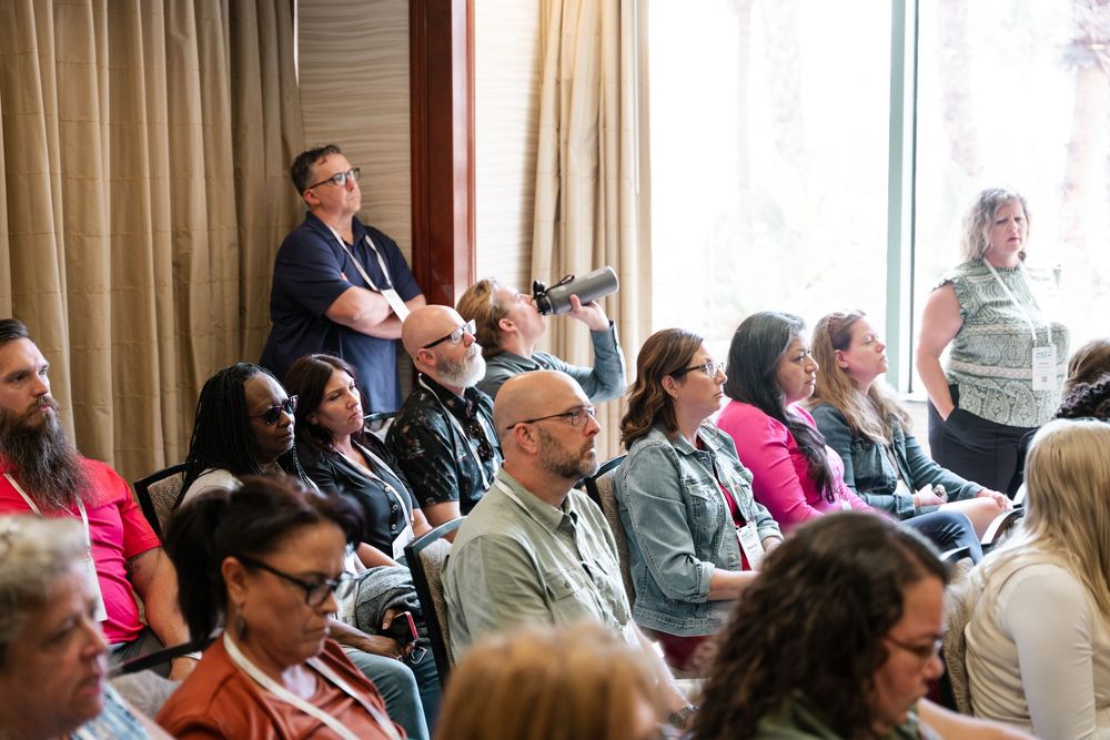 A large group of people seated in rows at a conference or meeting room with natural lighting from windows.