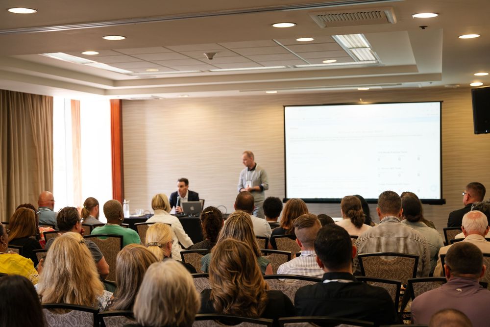 A speaker presents to an audience in a conference room with a projector screen and rows of attendees seated in chairs.