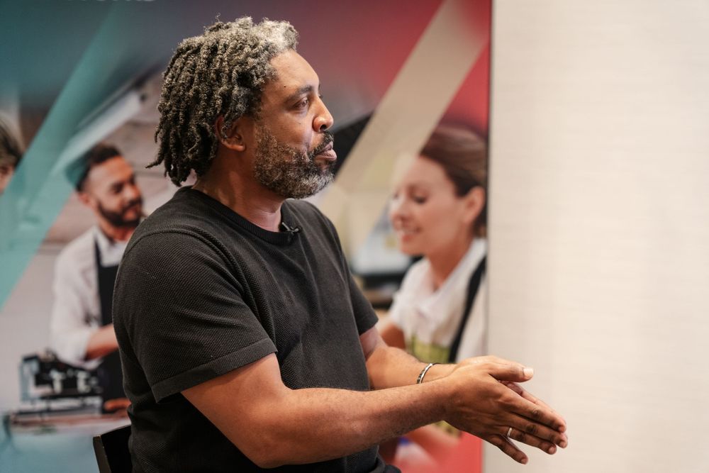 A person with curly hair demonstrates cooking techniques in a professional kitchen setting.
