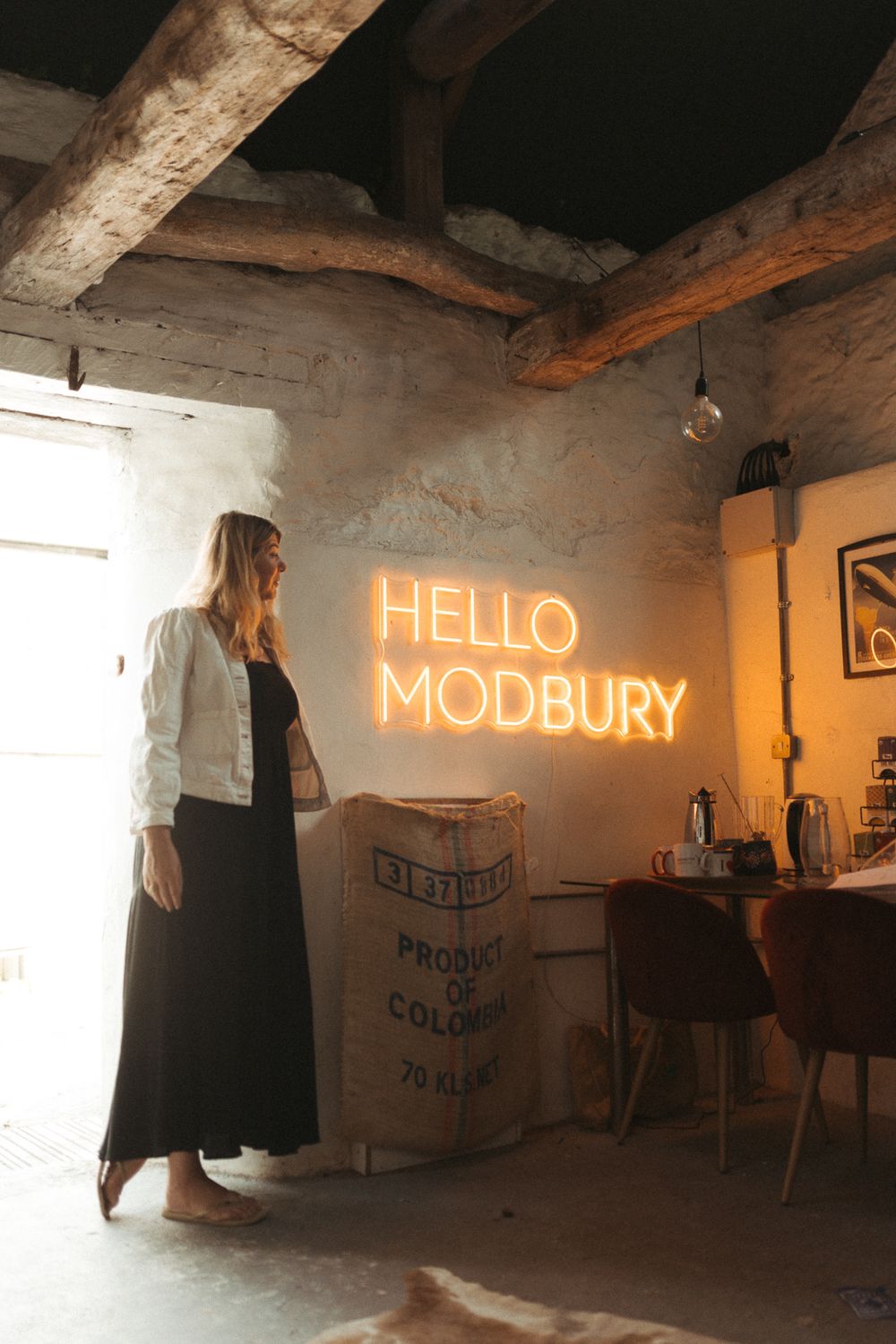 Interior of a rustic space with exposed beams and illuminated orange neon signs reading HELLO MODBURY.