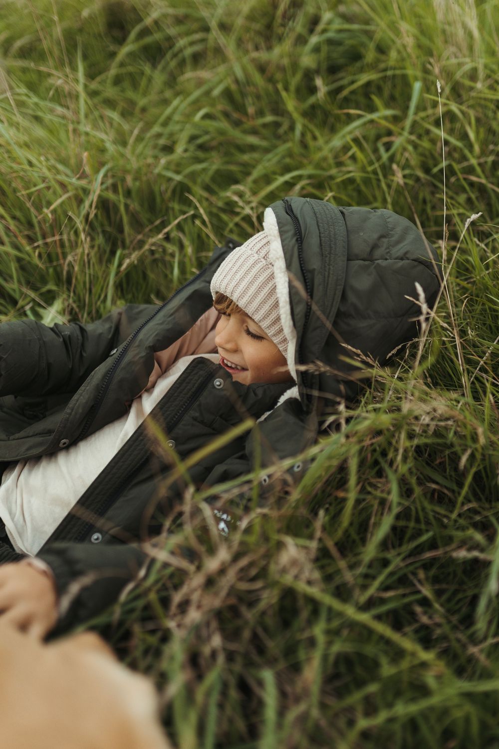 A child in a black puffer jacket and striped beanie lies smiling in tall green grass outdoors.