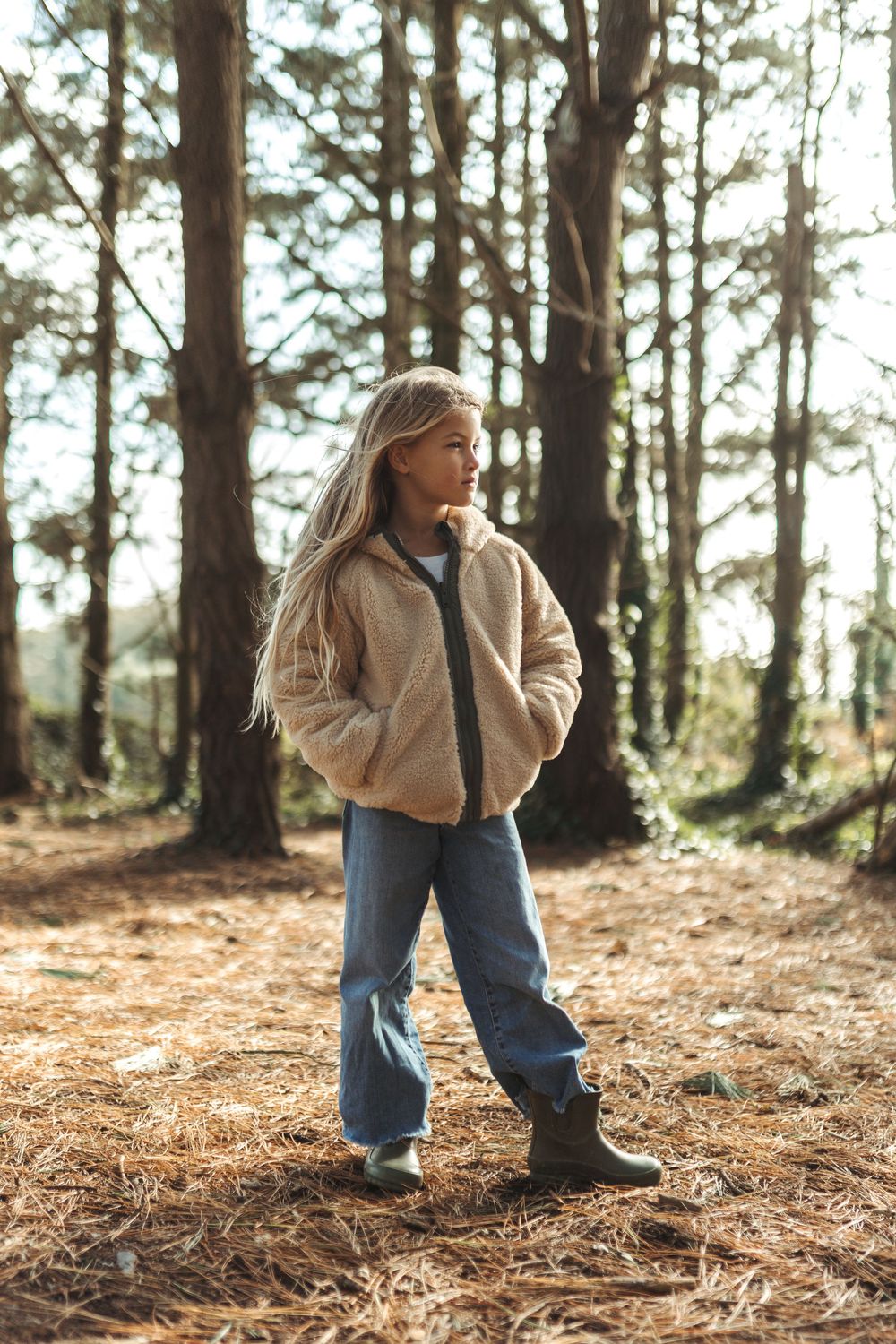 A young girl in a beige teddy jacket and jeans stands among tall pine trees in a sunlit forest.