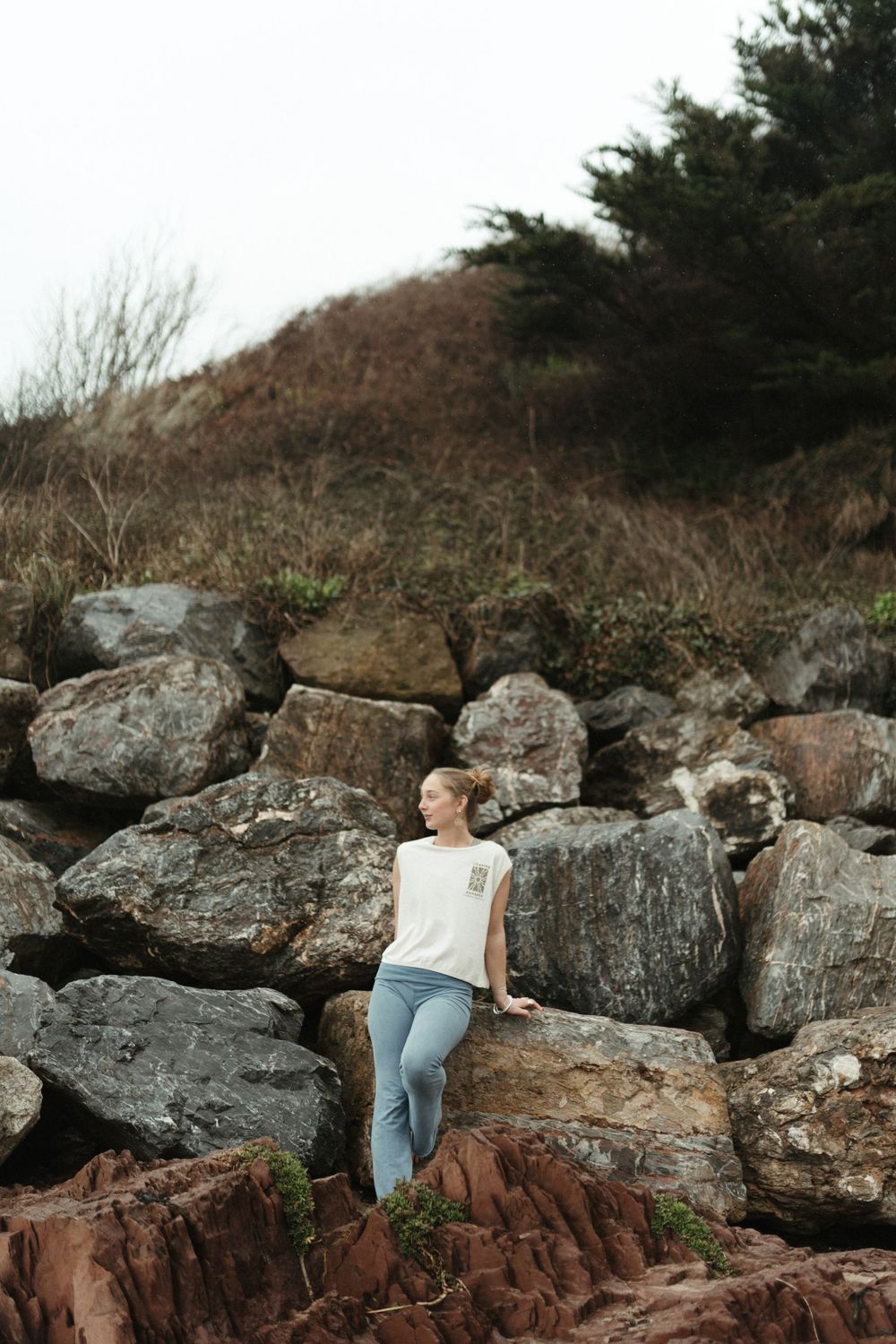 Woman in white top and blue jeans sitting on rocky coastal boulders with evergreen trees in background.