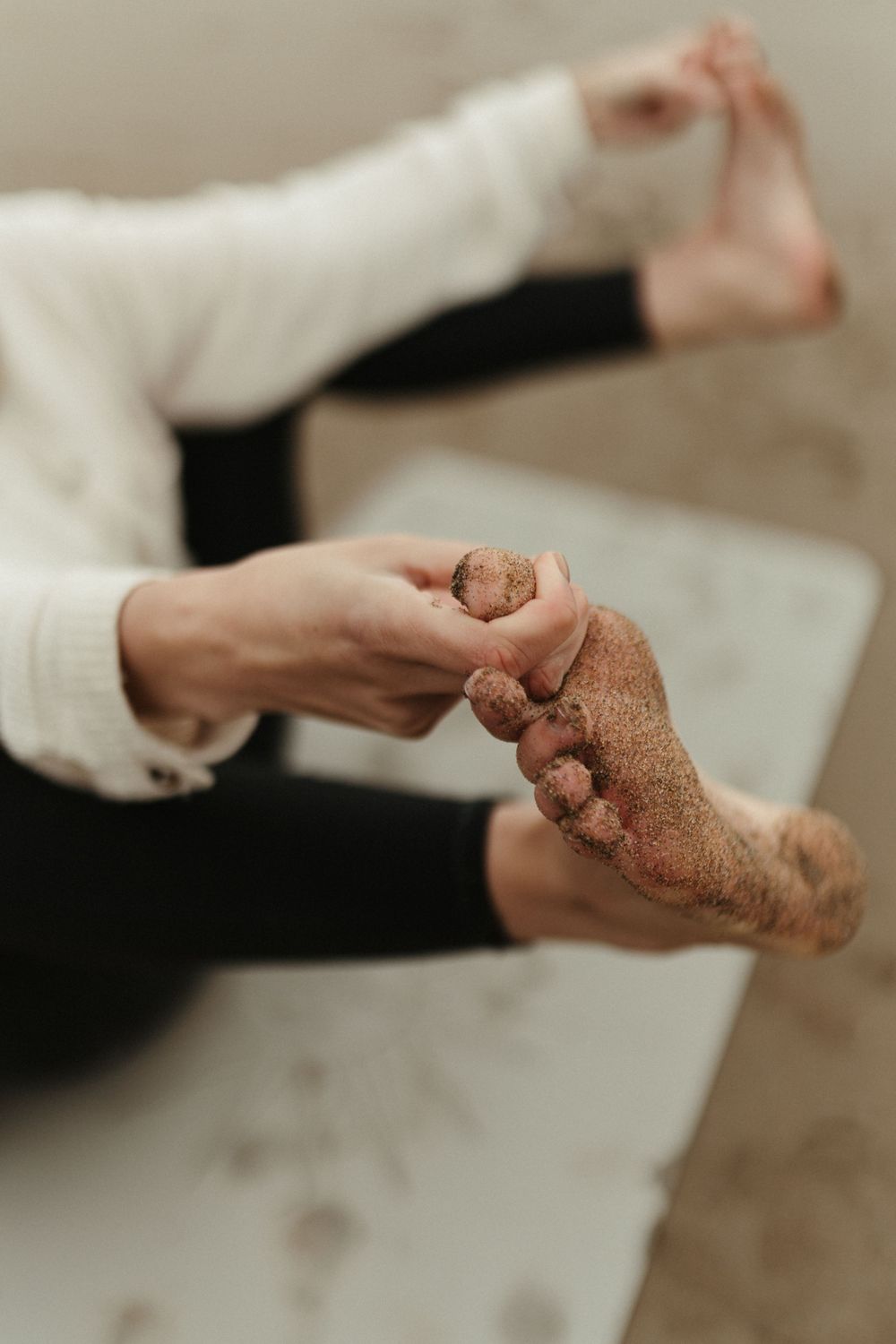 Close-up of sandy bare feet being massaged by hands wearing a white sweater indoors.
