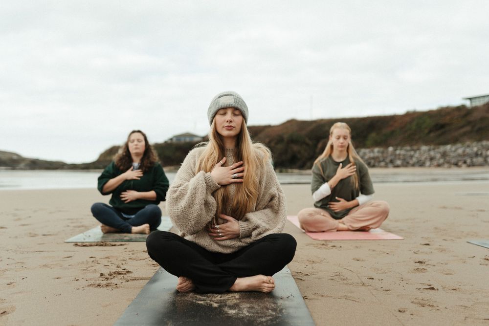 Three women meditating on yoga mats on a beach, sitting cross-legged with hands on hearts on a cloudy day.