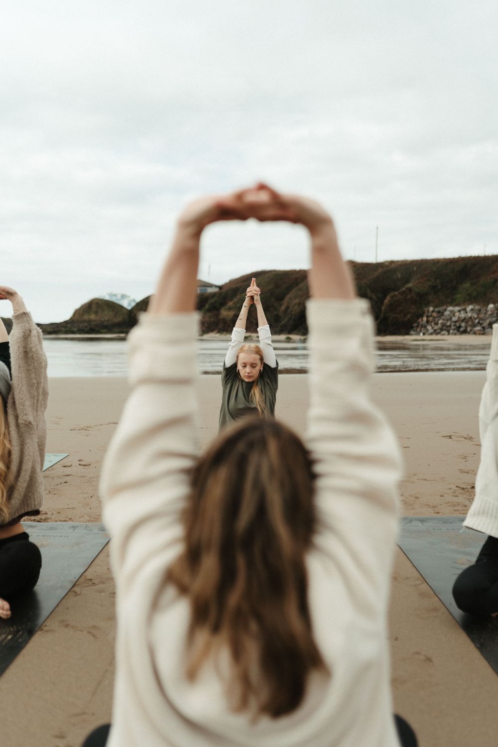 Group yoga class on a sandy beach with participants raising arms overhead, led by an instructor facing the ocean.
