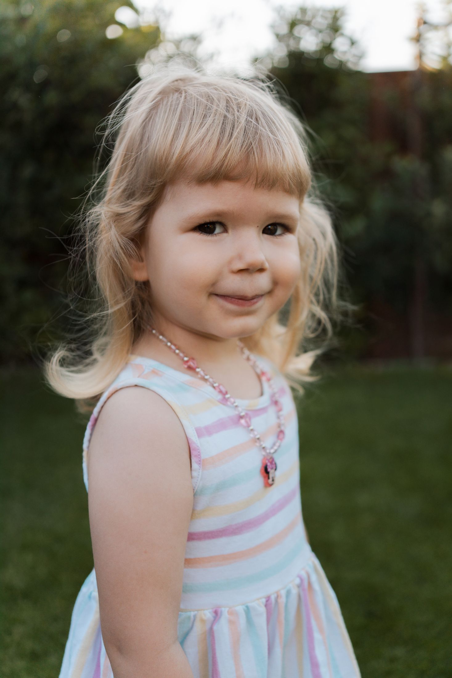 A child in a striped pastel dress smiles sweetly while playing outdoors at home in the garden in Rouse hill during a family photoshoot