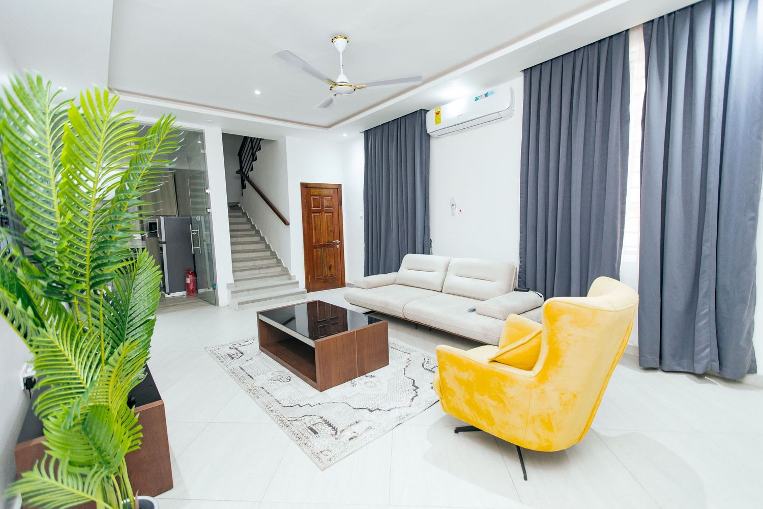 Contemporary white living space with wooden TV console, grey drapes, and tropical plant decor.