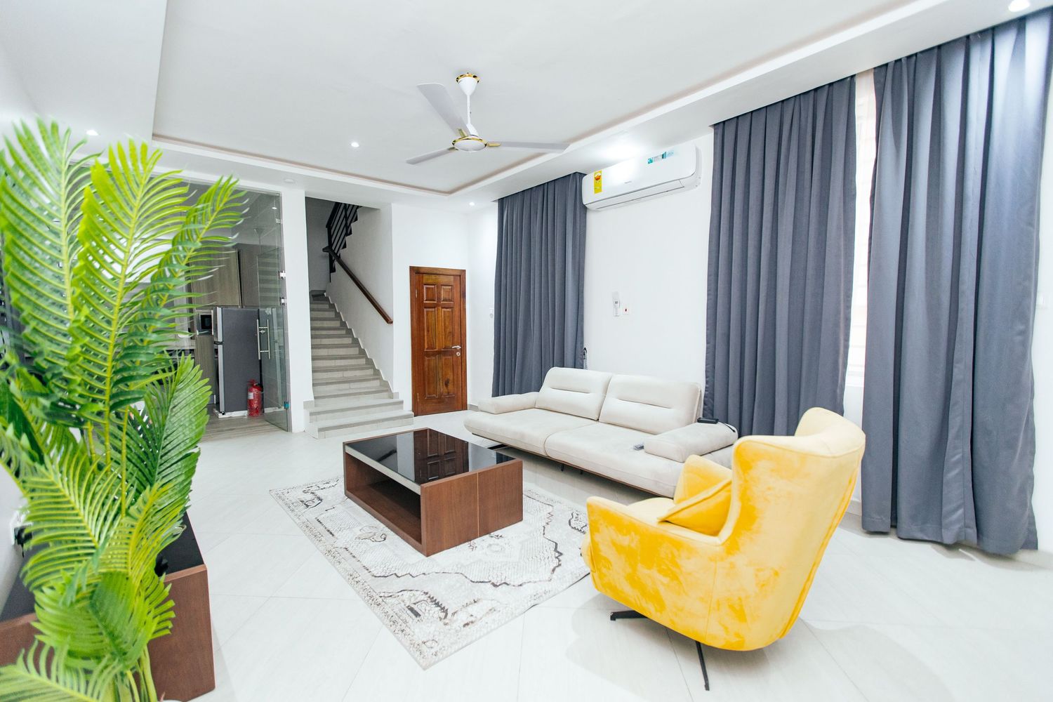 Modern minimalist living room with white walls, ceiling fan, and dark curtains features a bright yellow accent chair.