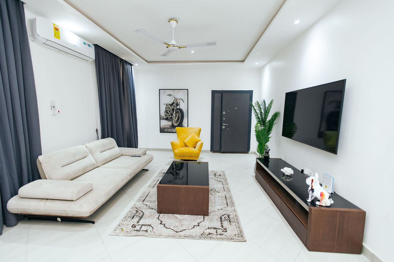 Contemporary living area featuring white walls, wall-mounted TV, and comfortable seating with dark window treatments.