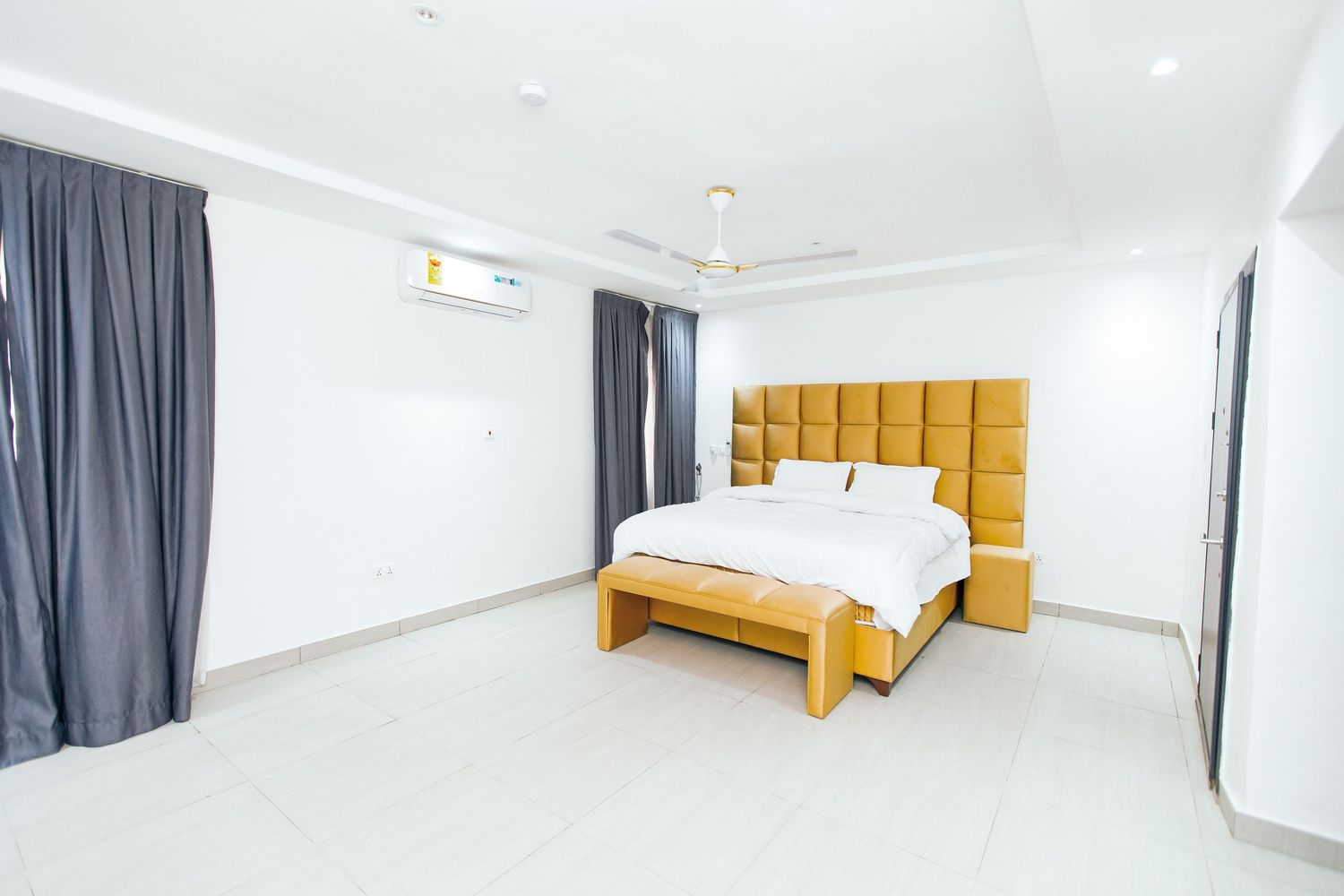 Spacious bedroom with mustard yellow headboard, gray curtains, and bright white walls.