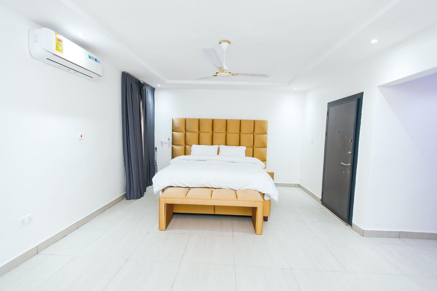 Modern bedroom interior with yellow tufted headboard and dark curtains against white walls.