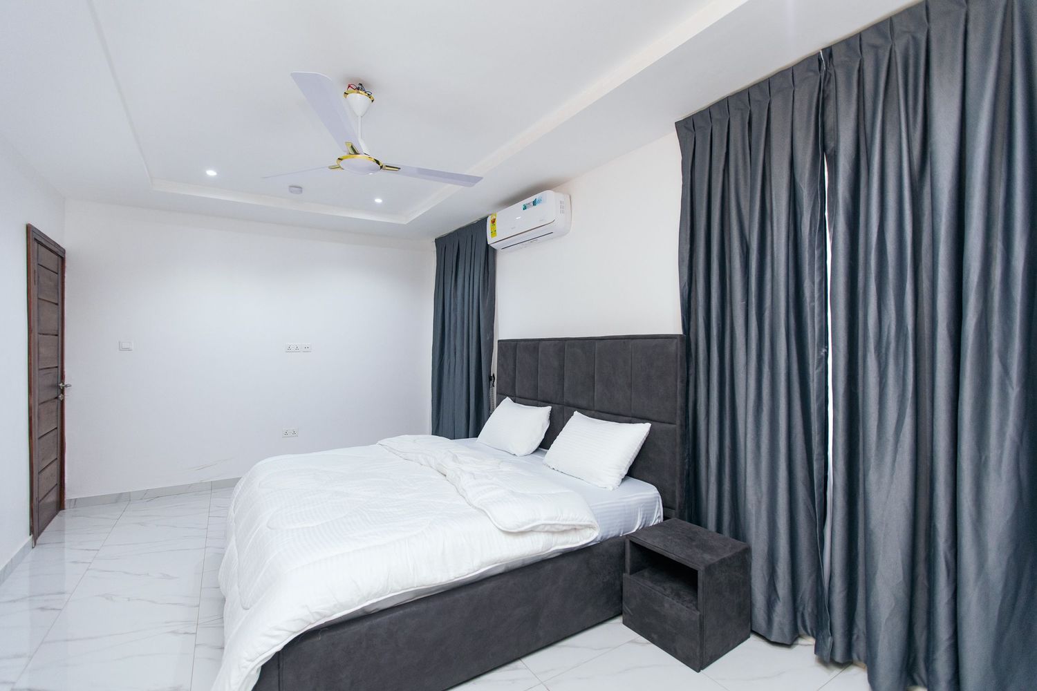 Modern bedroom featuring grey curtains, white walls, and comfortable bedding setup.
