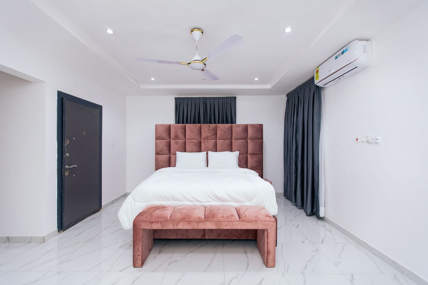 Spacious white bedroom with mounted TV, teal curtains and pink velvet headboard.