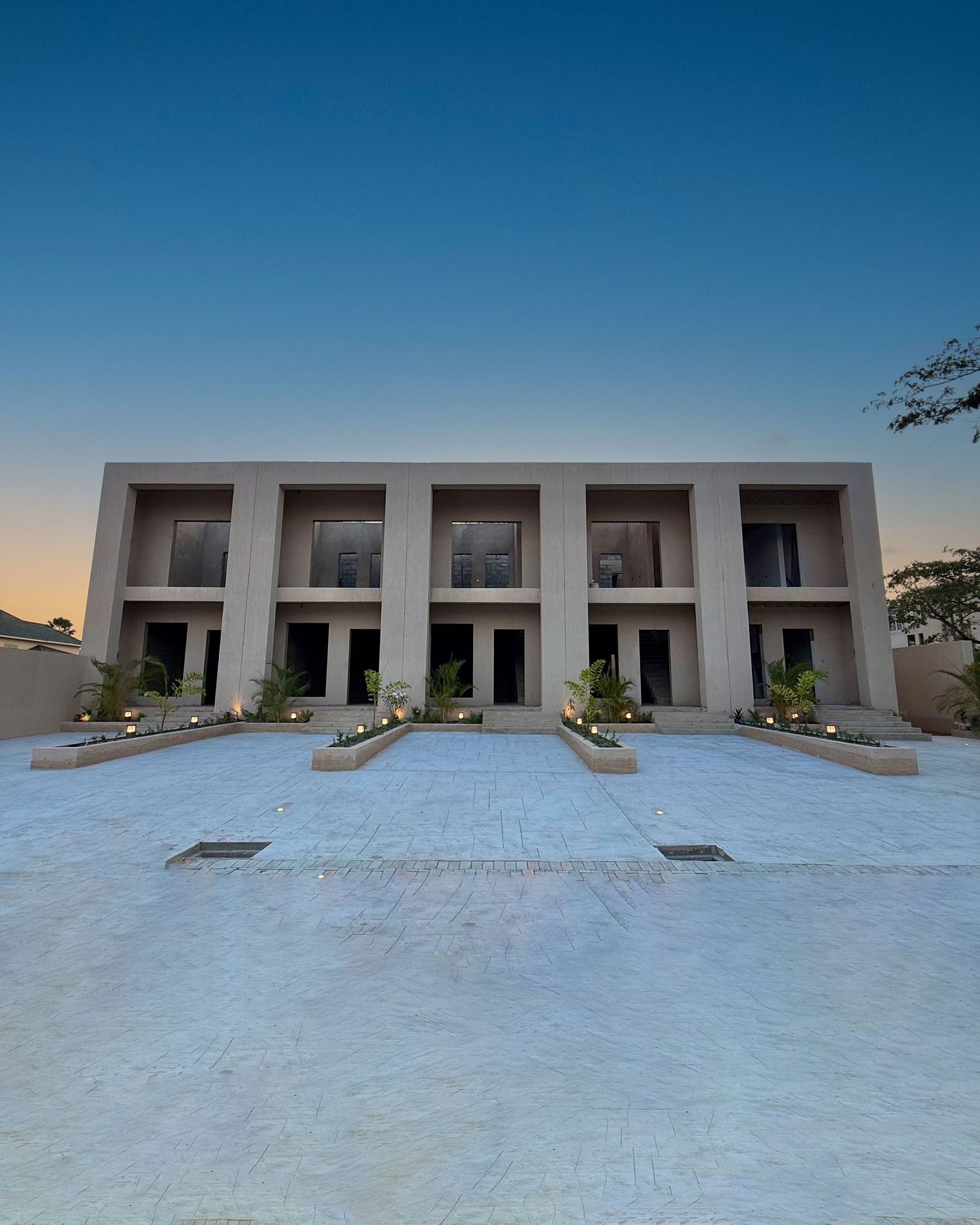Modern minimalist townhouse building with five identical units featuring white walls and glass doors on a concrete plaza at dusk.