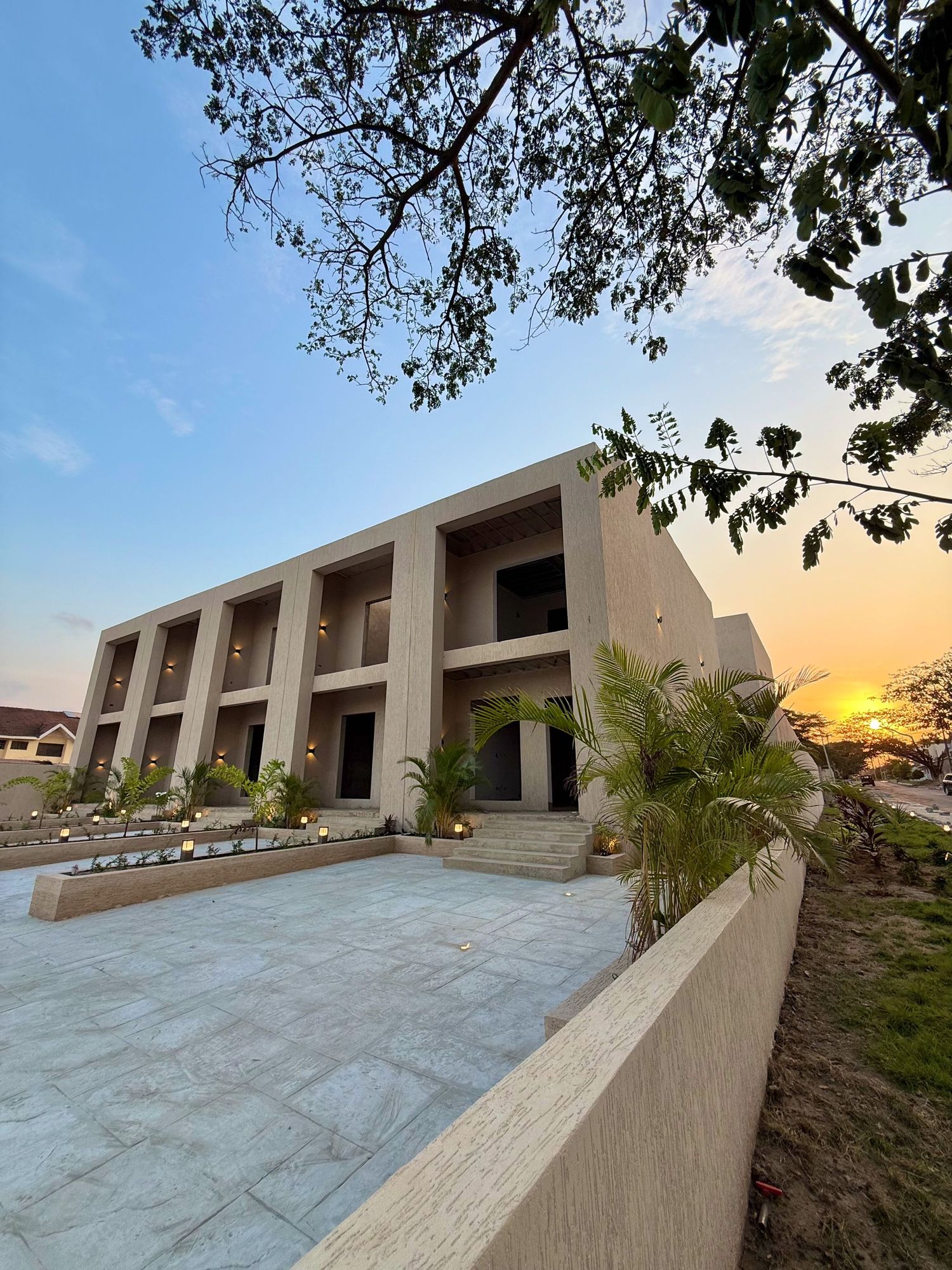Modern architectural design of a beige building with geometric shapes and palm trees against a sunset sky.