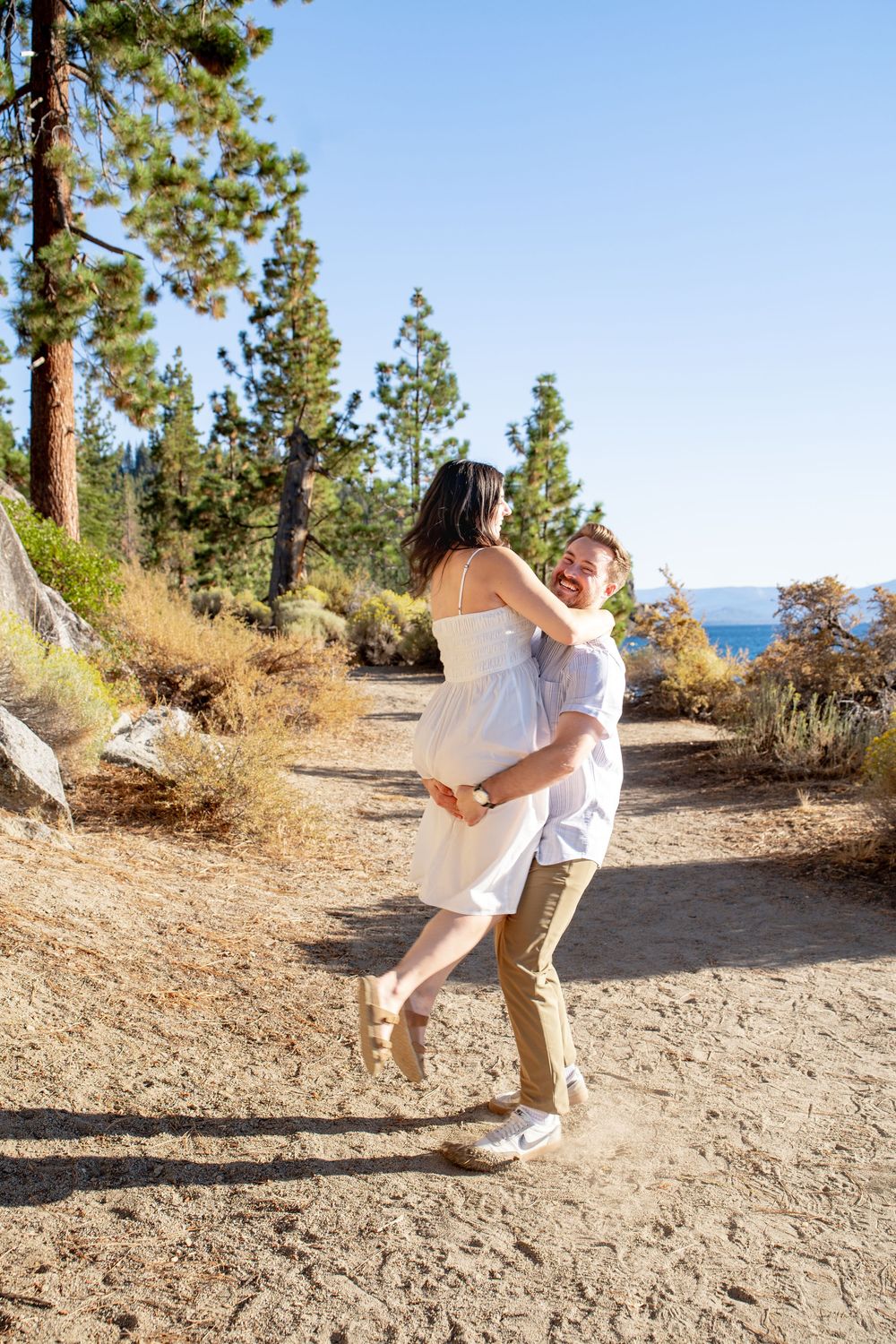 A couple dances and twirls playfully on a scenic mountain trail overlooking a lake on a sunny day.